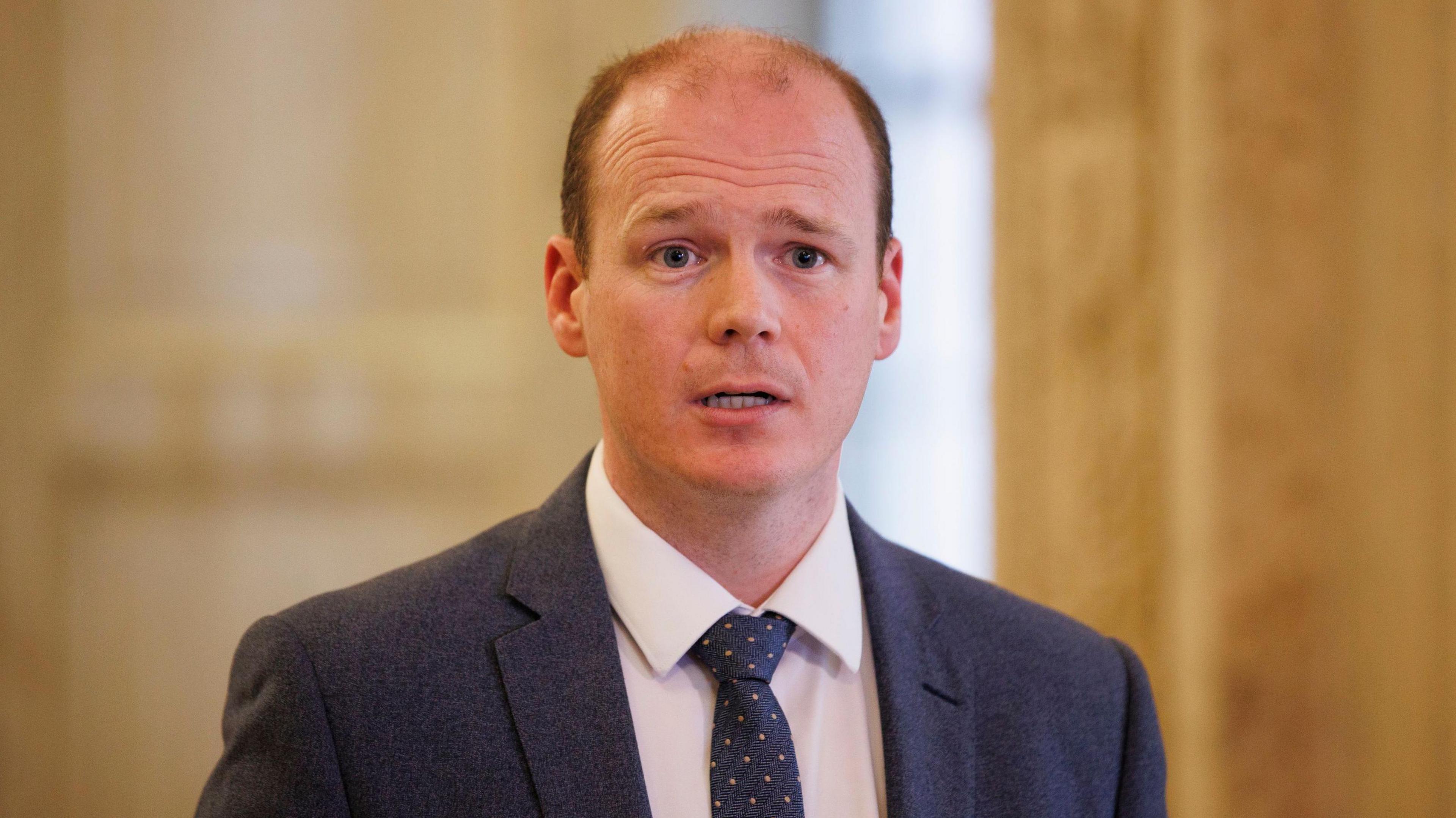 Gordon Lyons speaks in Stormont's Great Hall. He is stood against a white marble wall. He is a balding man with brown hair, and he is mid-speech. He is wearing a dark grey suit jacket, a white collared shirt and a dark tie.