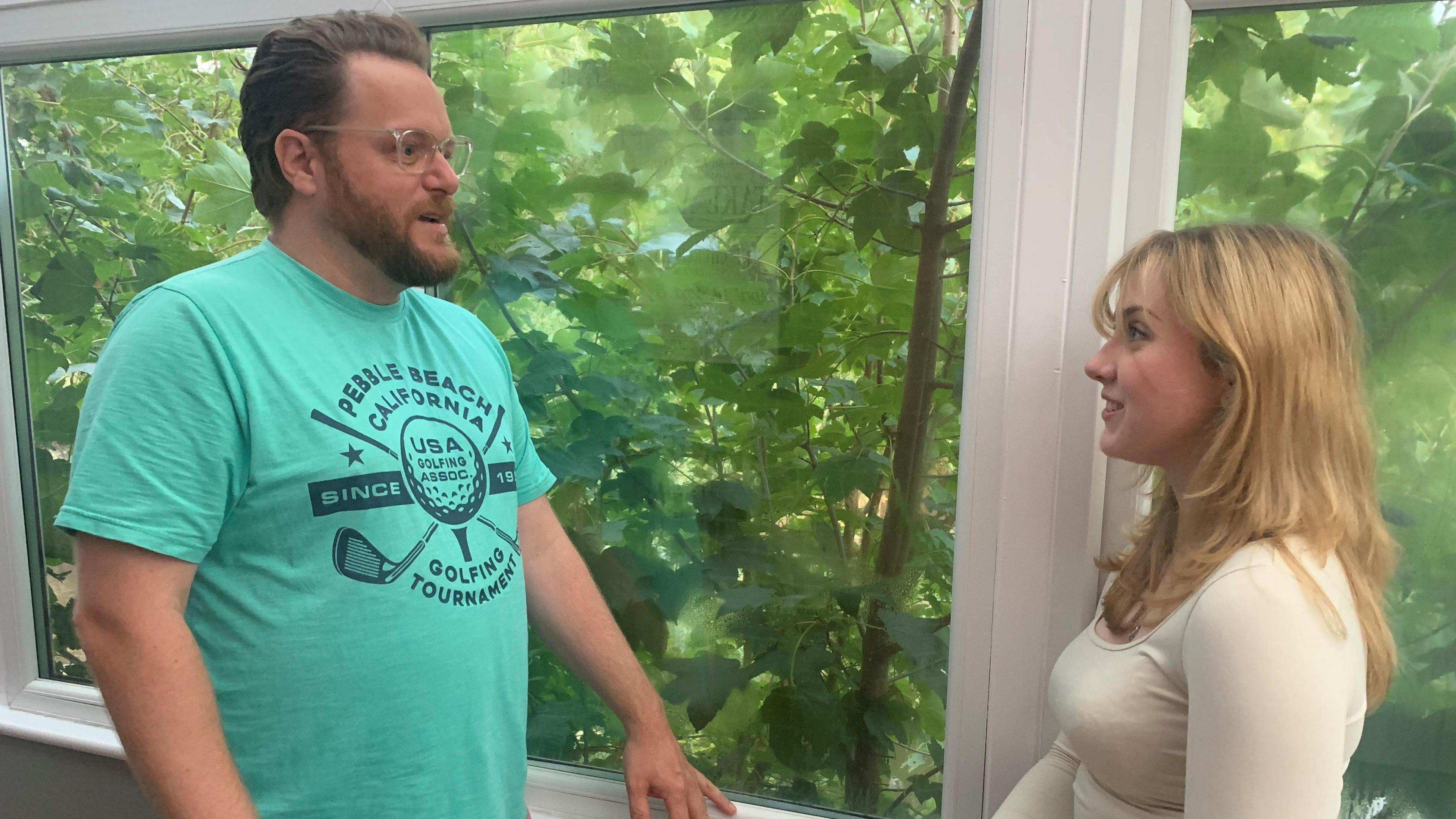 A man wearing glasses and a green T-shirt speaking to a woman with blonde hair wearing a white top next to a window with trees outside