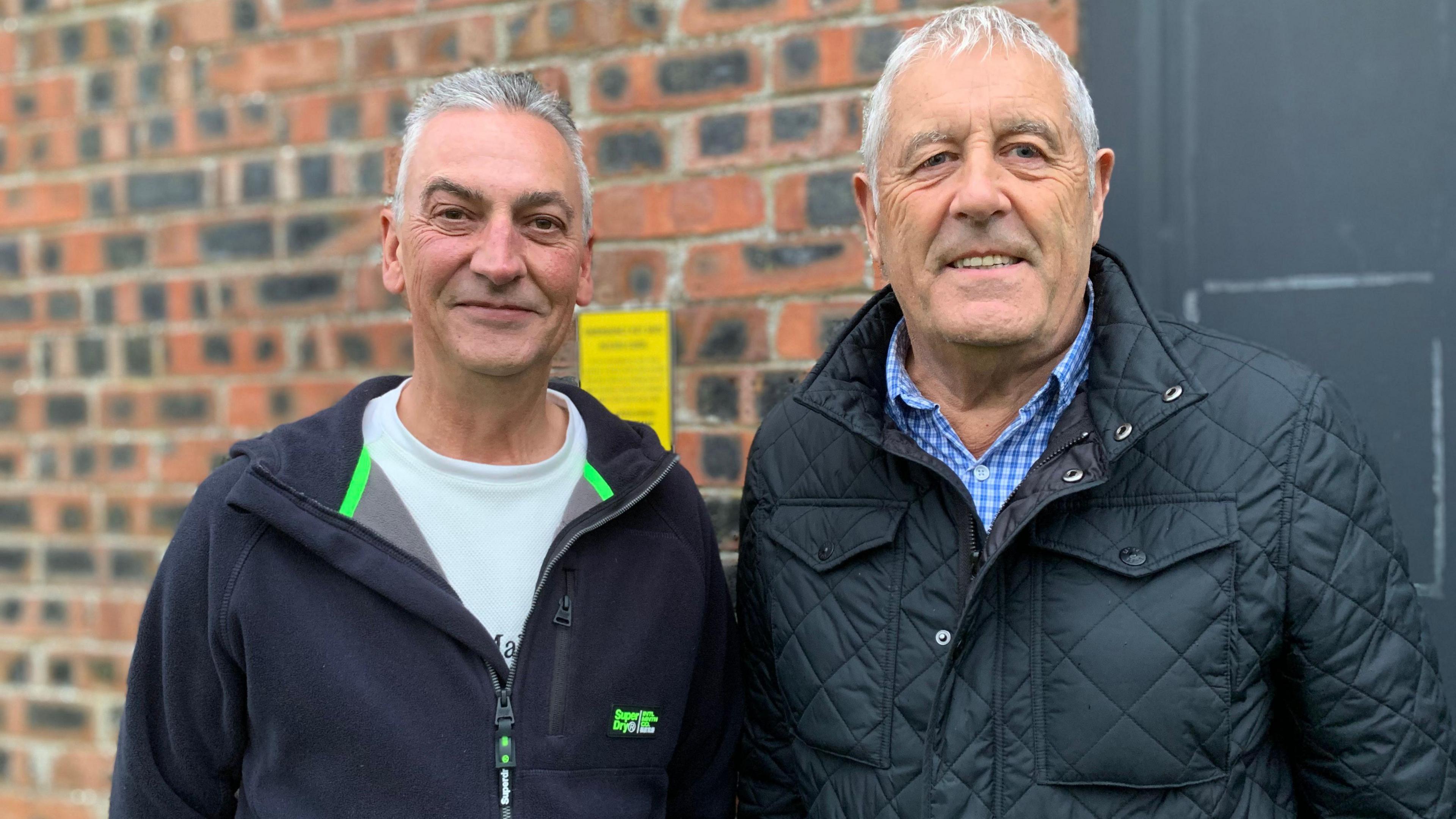 Two men with grey hair stand in front of a brick wall. One is wearing a black zip top with a white T-shirt underneath and the other is in a padded black coat with a blue checked shirt.