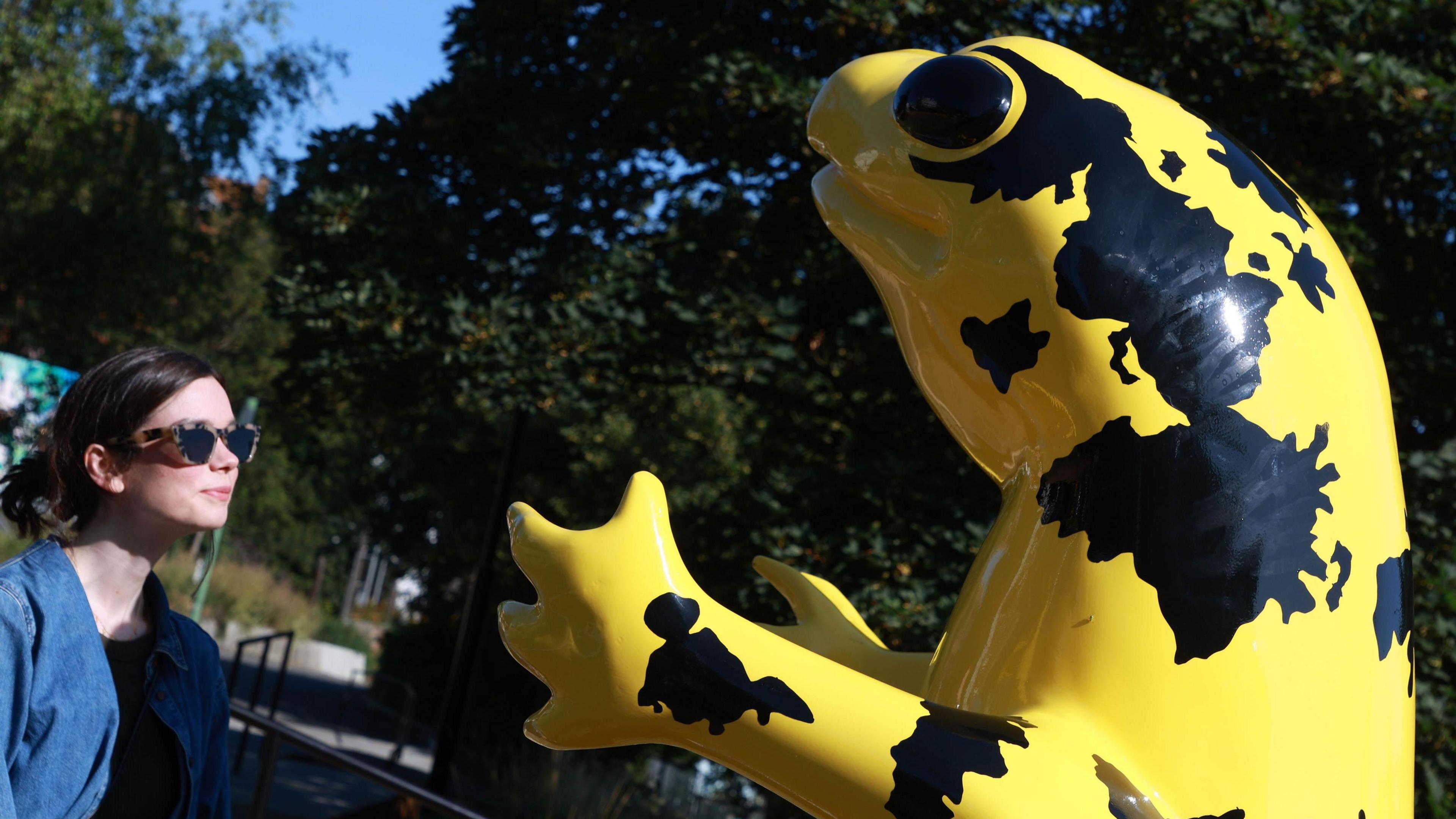 A large statue of a yellow and black frog-like creature with a woman in sunglasses looking at it