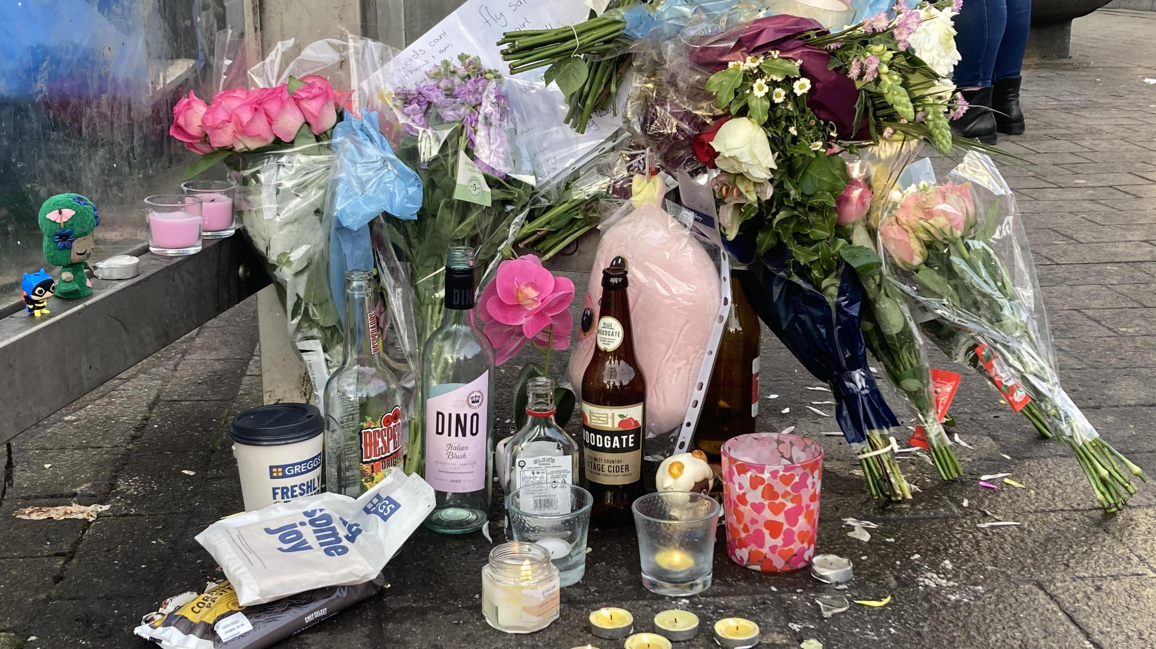 A pile of bunches of flowers, cards, empty bottles, lit candles, and other food items and trinkets at what appears to be the base of a bus stop.