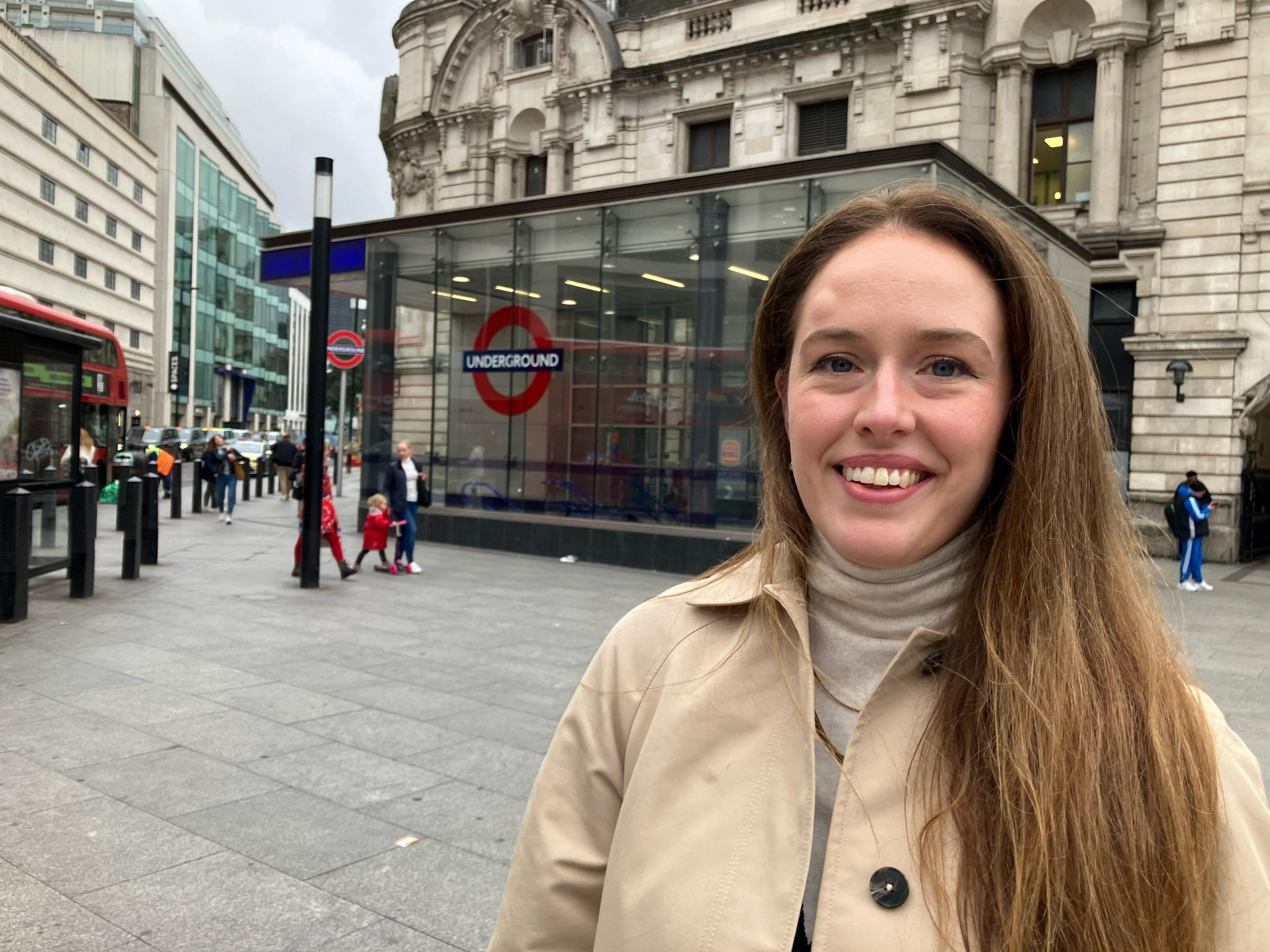 Franki Oliver from Royal National Institute for Deaf People smiling to camera with long brown hair and wearing a beige raincoat. Behind her is an entrance to the Tube outside Victoria station with a red double decker bus passing in the distance.