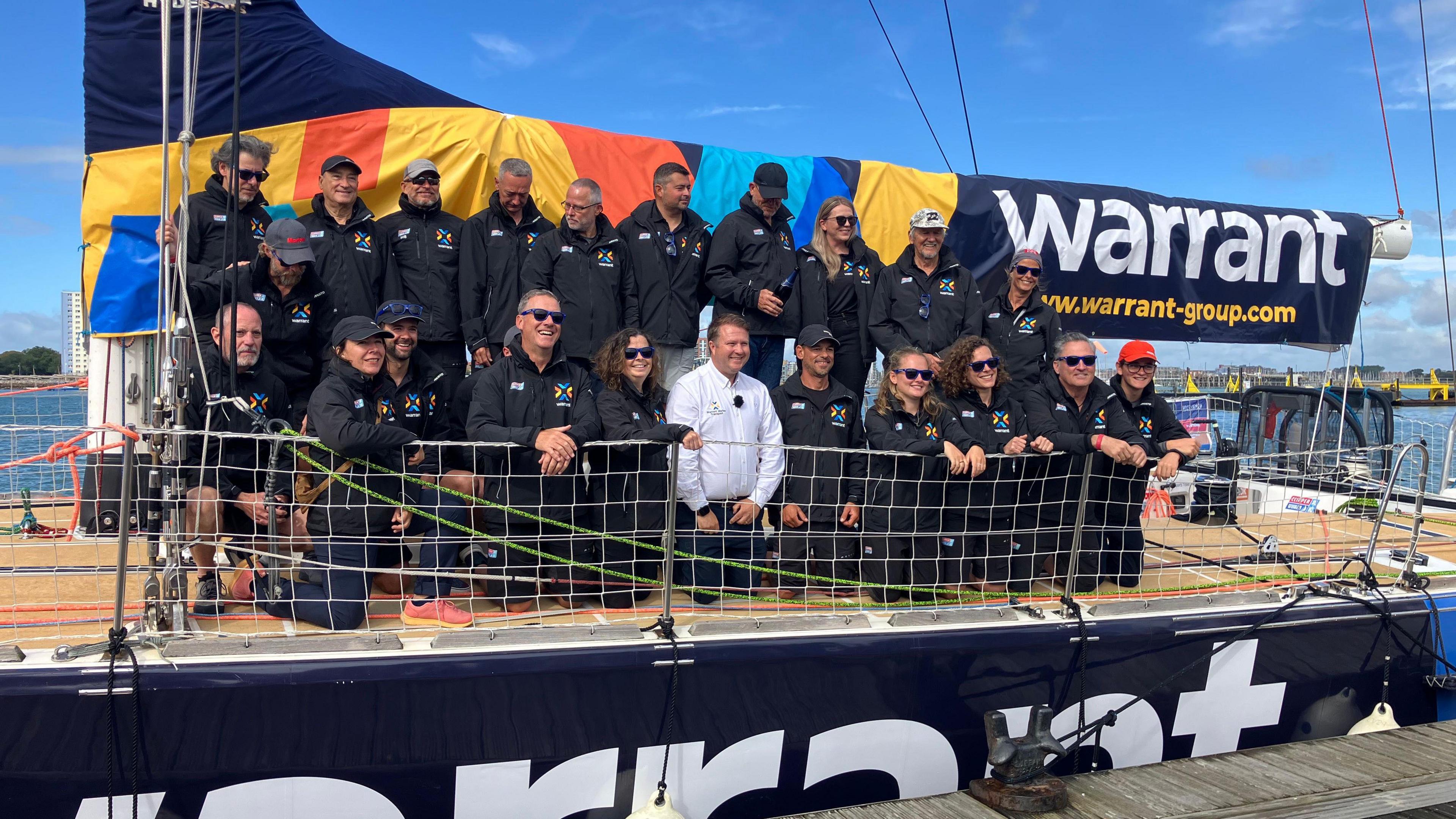 A large number of people sitting and kneeling in two rows on a boat deck. All the people, other than a man kneeling in the centre of the front row are wearing branded jackets. Behind them, the sail has the word "Warrant" and an email address written prominently on it.