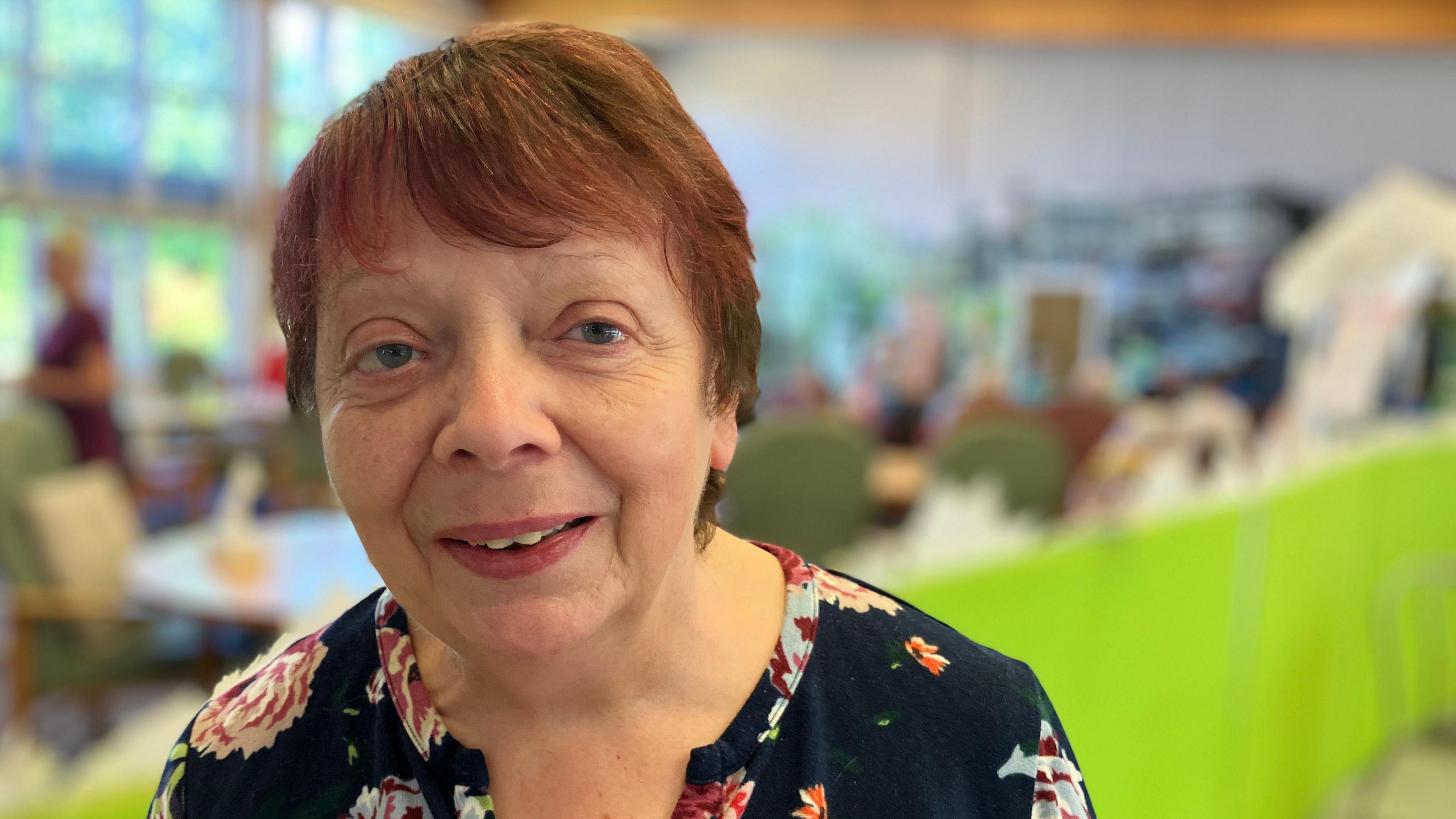 A close-up of manager Eileen Wright, who has short, dark red hair and wears a dark top with a floral pattern. She stands inside the Endike Community Care centre.