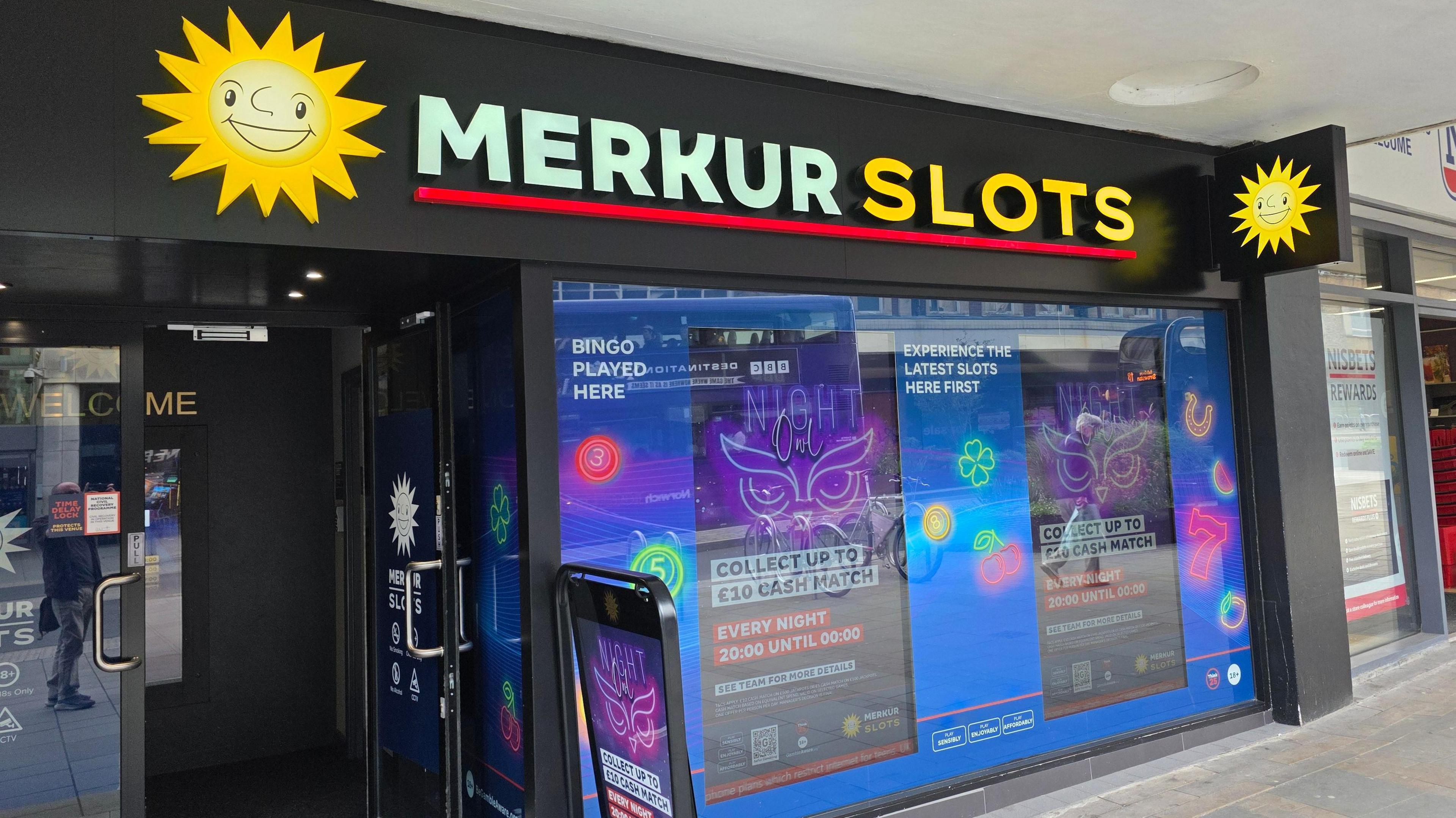 We can see the exterior of a Merkur Slots branch. At the top is the shop name, with two shining 'smiley face' suns next to it. In the shop window in a gambling offer.