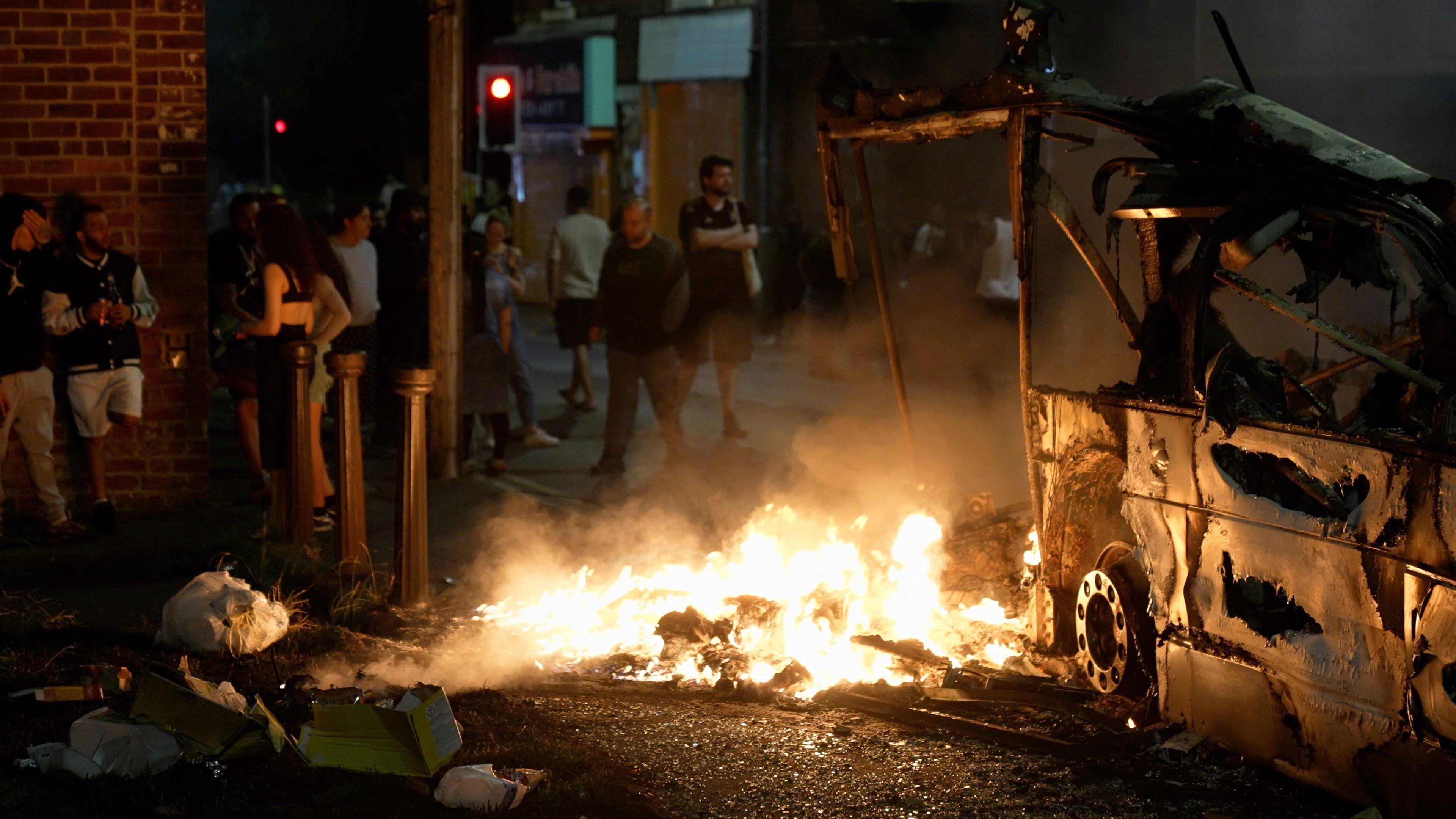 The carcass of a vehicle is on fire at night. Crowds gather around.