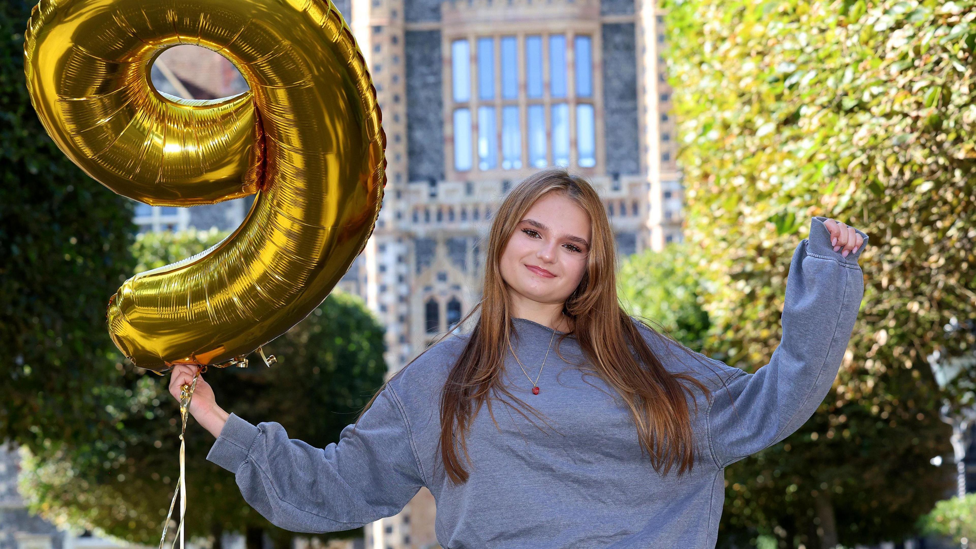 A teenager wearing a light blue top and holding a large gold balloon in the shape of the number 9. She is holding her hands in the air and smiling.