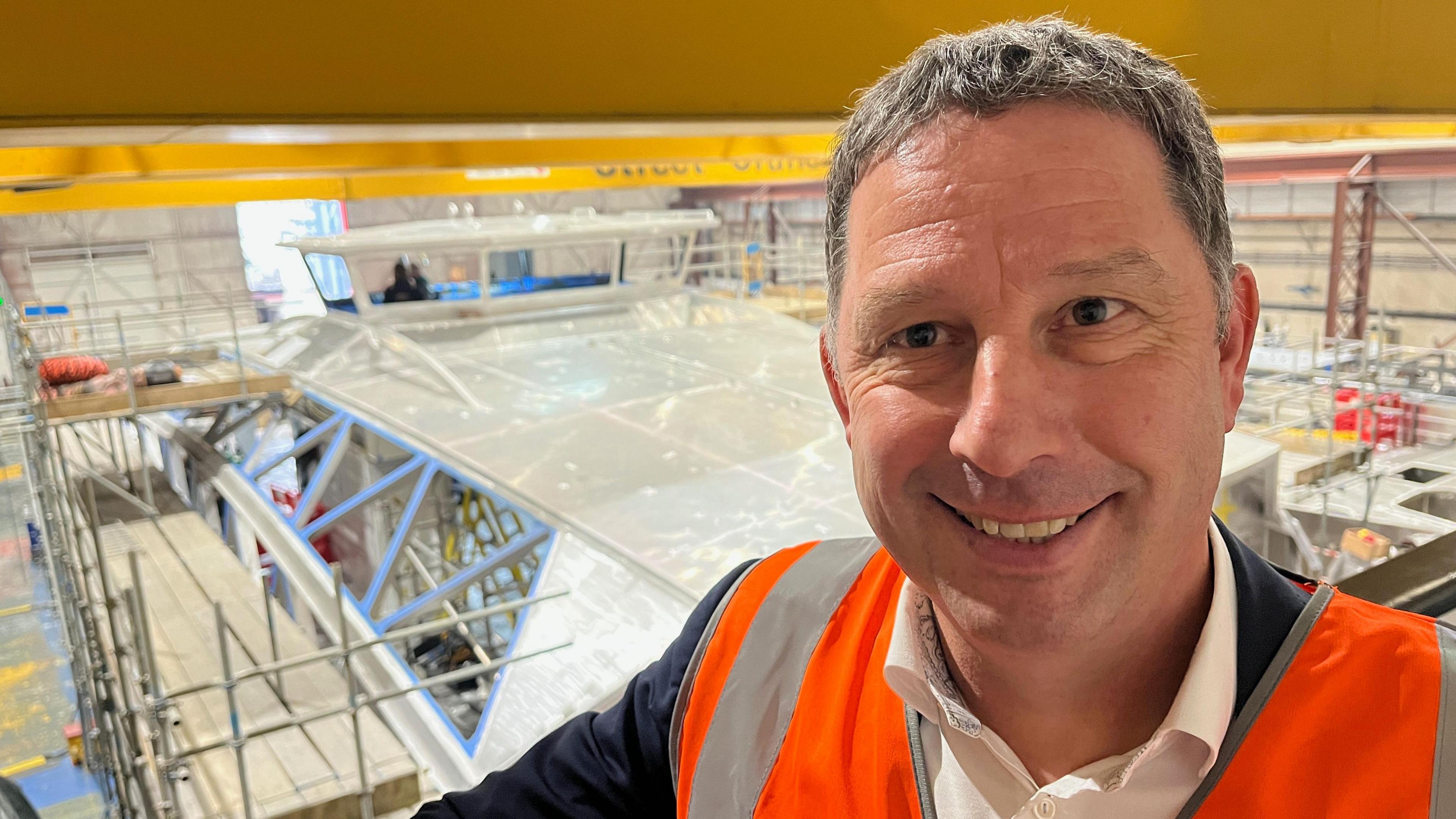 Sean Collins has short grey hair and is wearing an orange hi-vis jacket over a white shirt and blue blazer. Behind him is the metallic shell of the new ferry