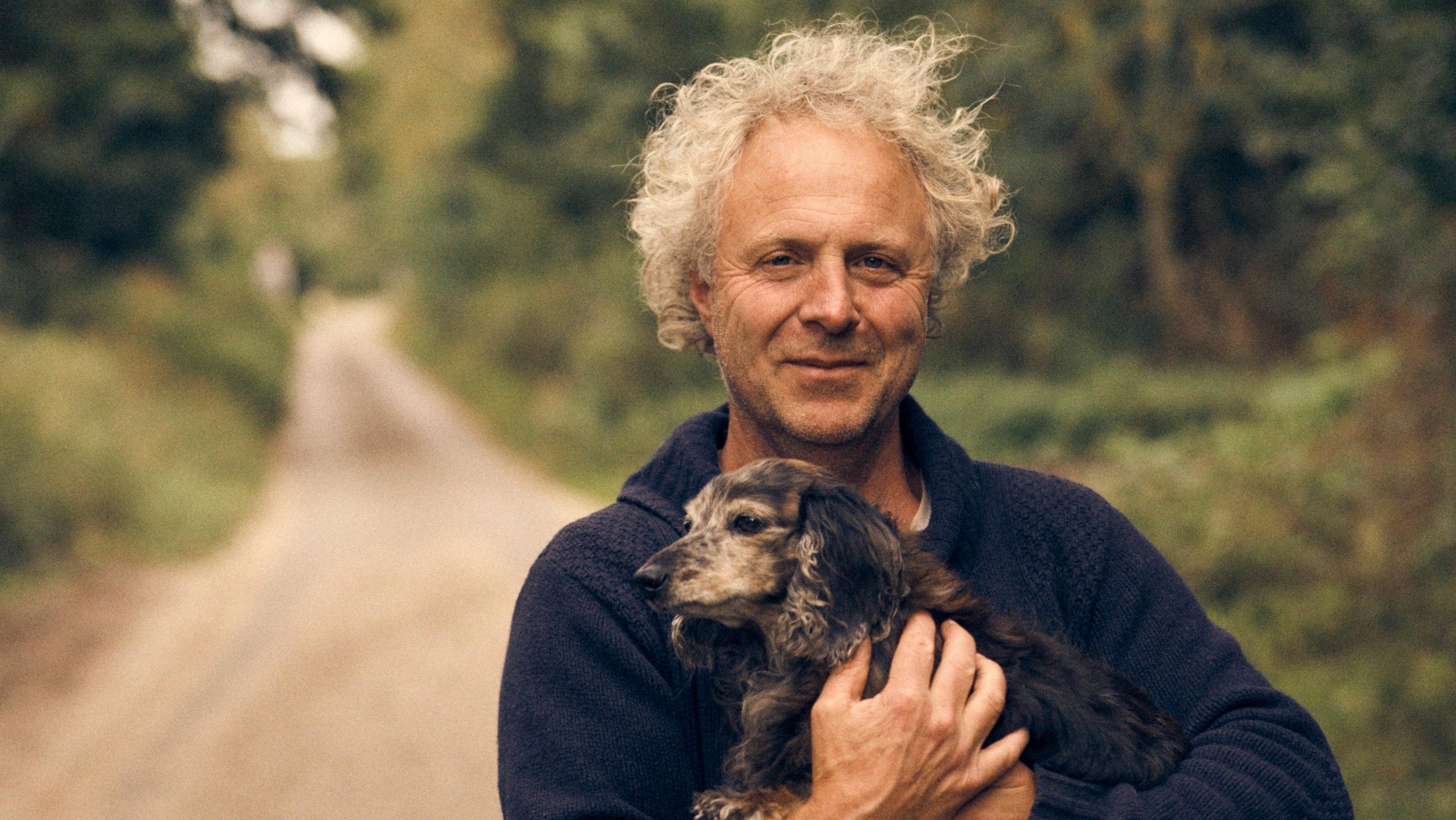 Charlie Mackesy, with a country lane behind him, smiles as he holds his little dog Barney in his arms