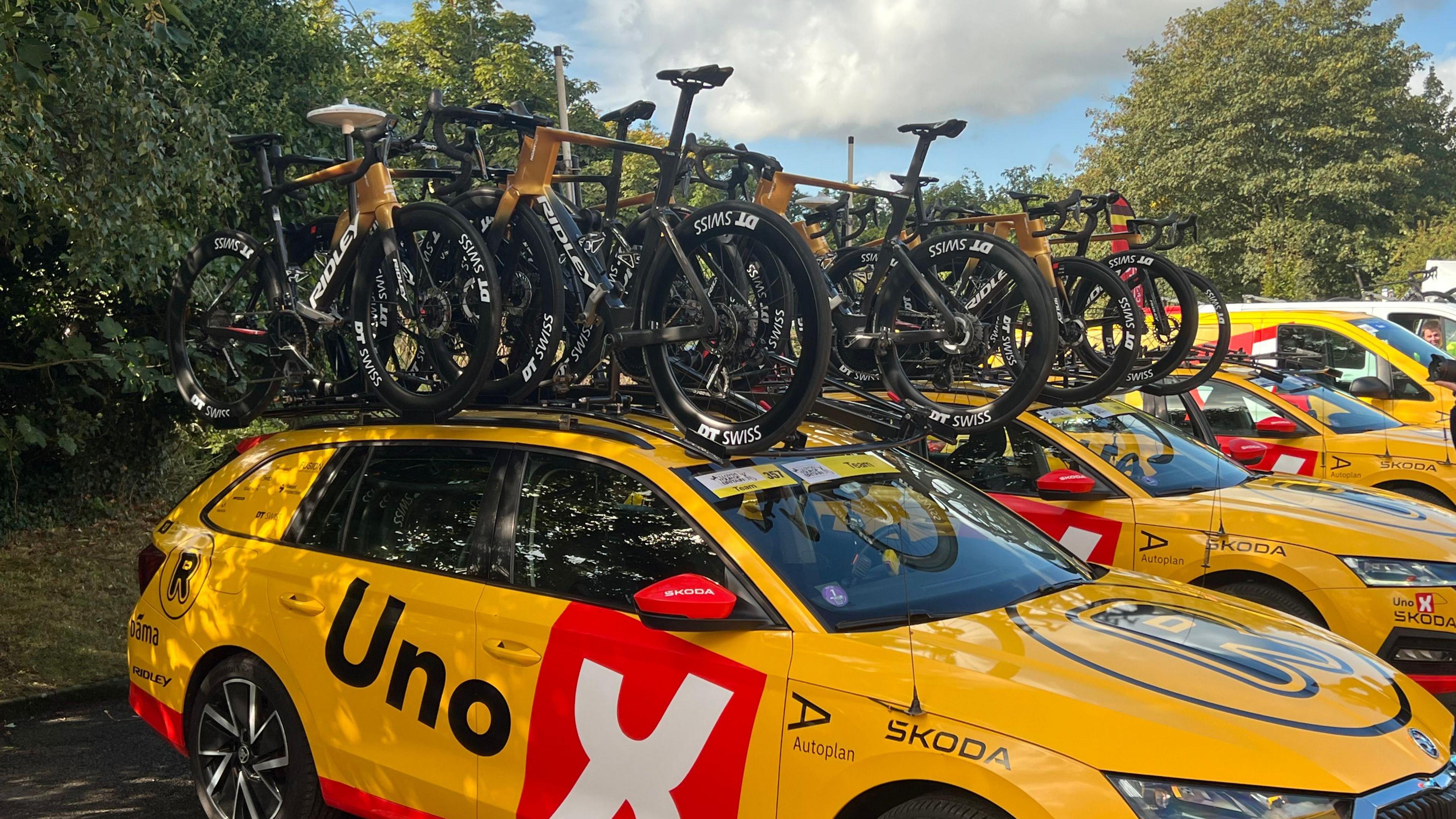 Stacks of black and gold racing bikes sit in a rack on top of a yellow car that has various branding painted on to it. 