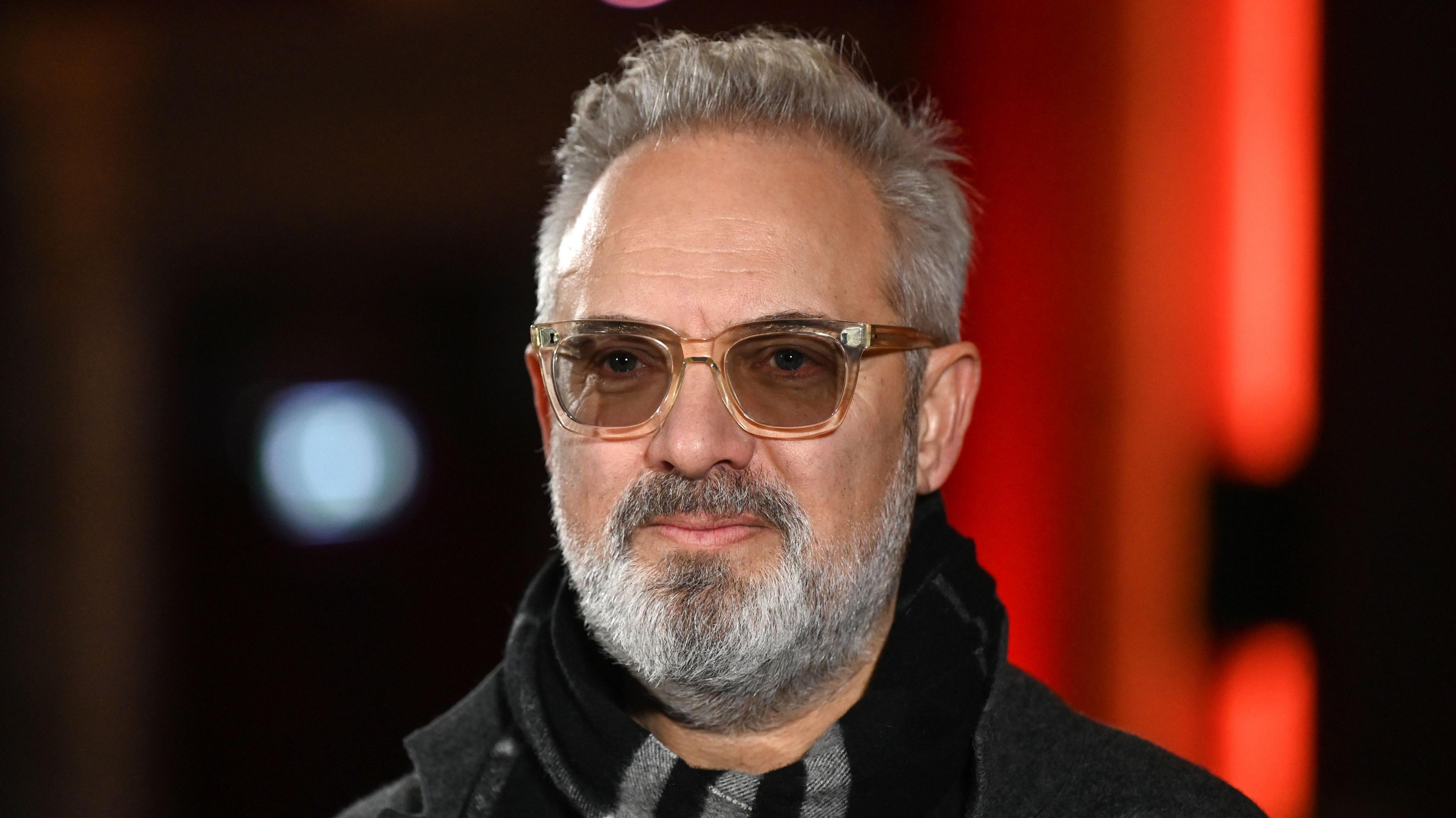 Sam Mendes smiling at the camera with brown framed glasses and grey facial hair.