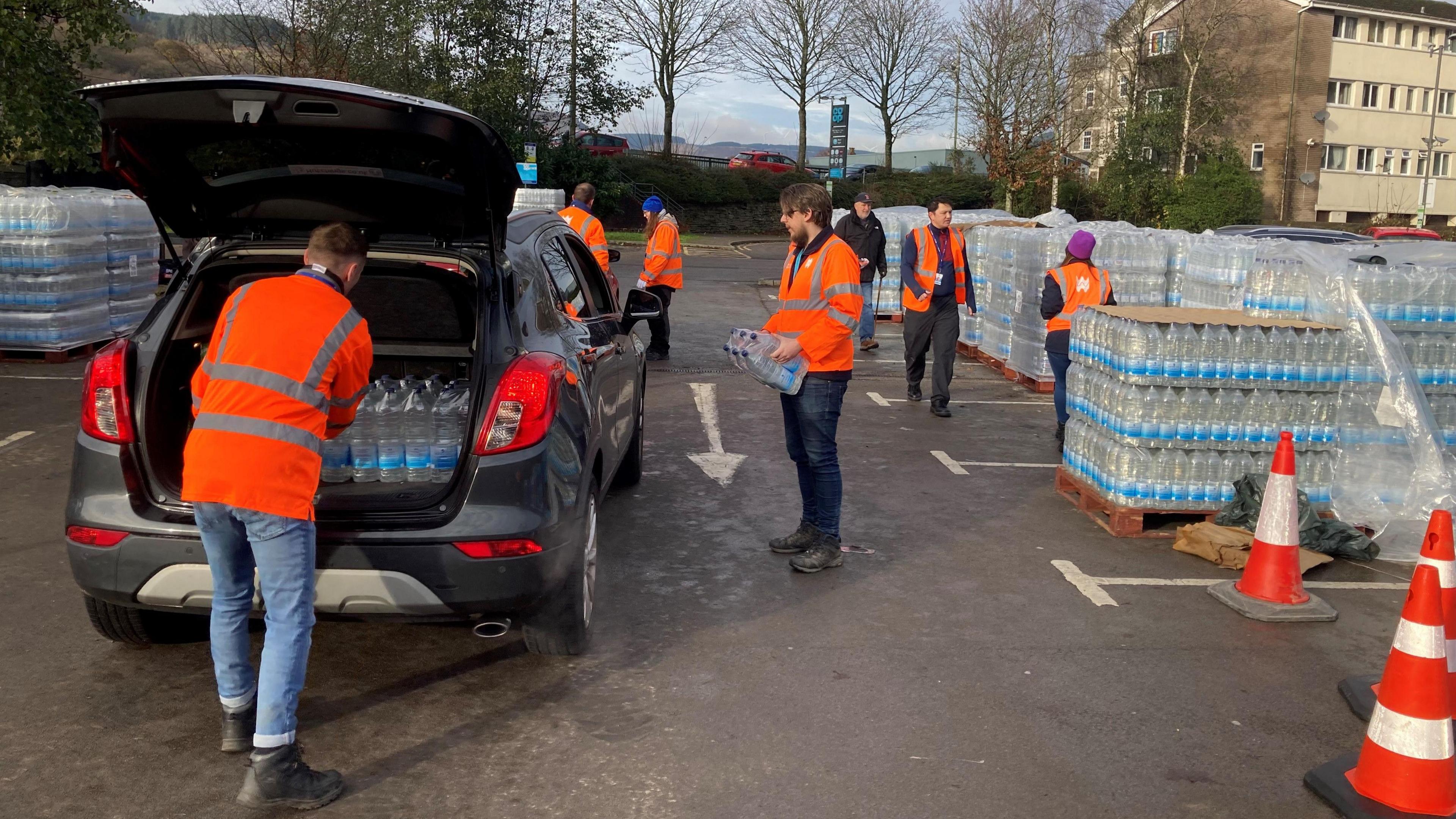 Welsh Water staff in orange high-vis jackets fill the boot of a car with bottled water at one of their bottled water stations, situated in the Co-op car park in Treorchy.