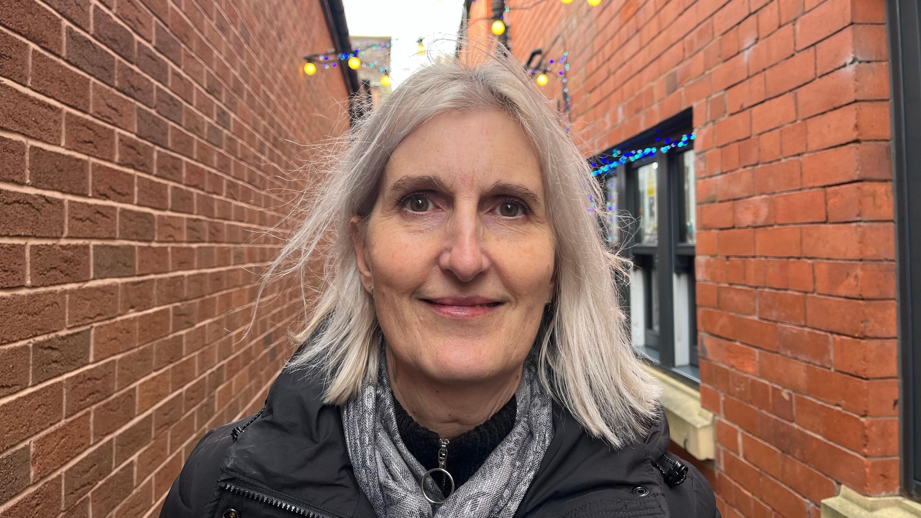 Adele looking into the camera, she's wearing a black jumper, a grey scarf, and a black winter coat. She's stood in one of Oswestry's side streets, you can see Christmas lights above her head and a shop window to her left.