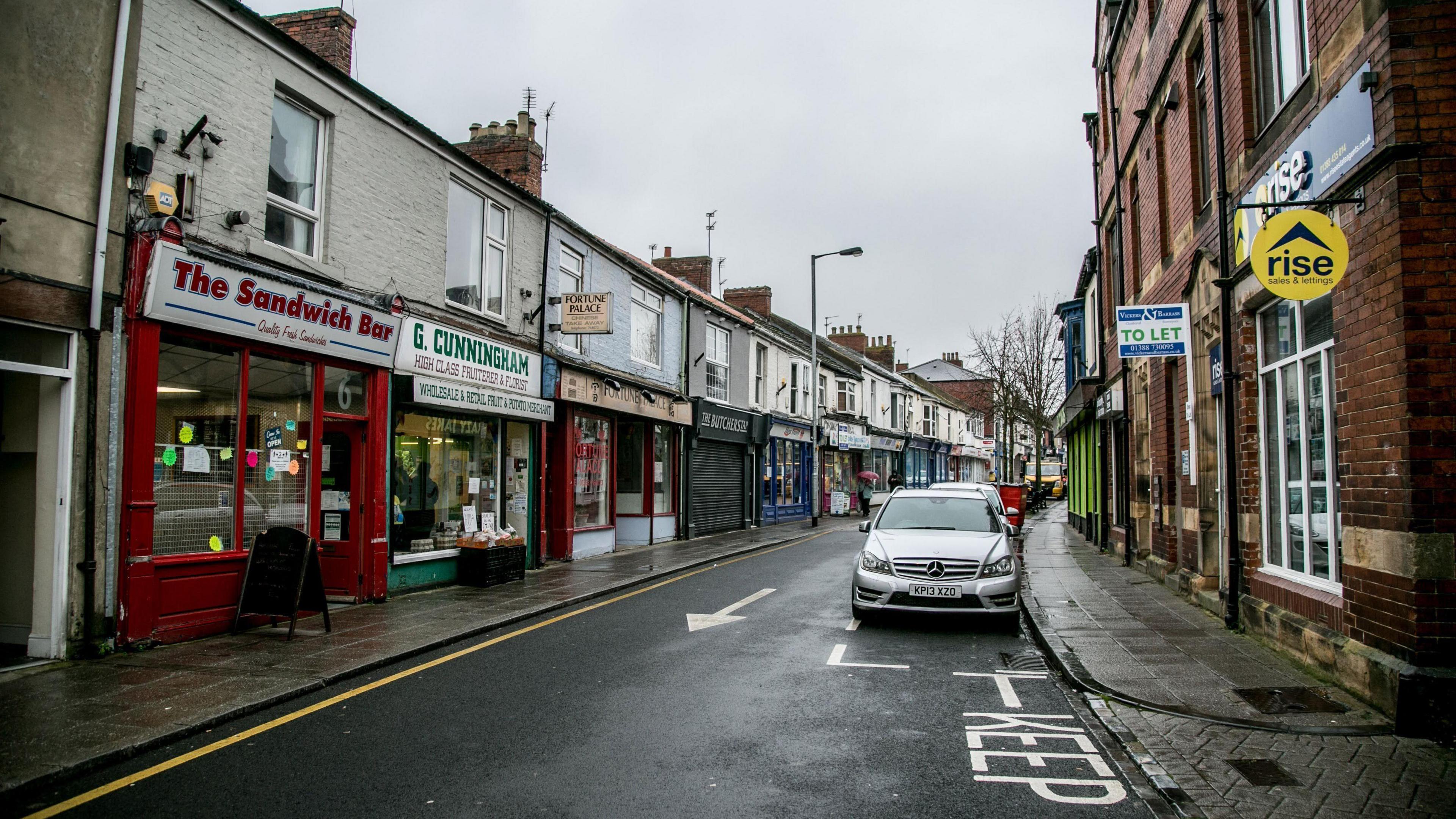 Crook town centre. A sandwich shop is one of many shops lining a high street made up of a row of terraced buildings. Cars are parked on the other side of the road.