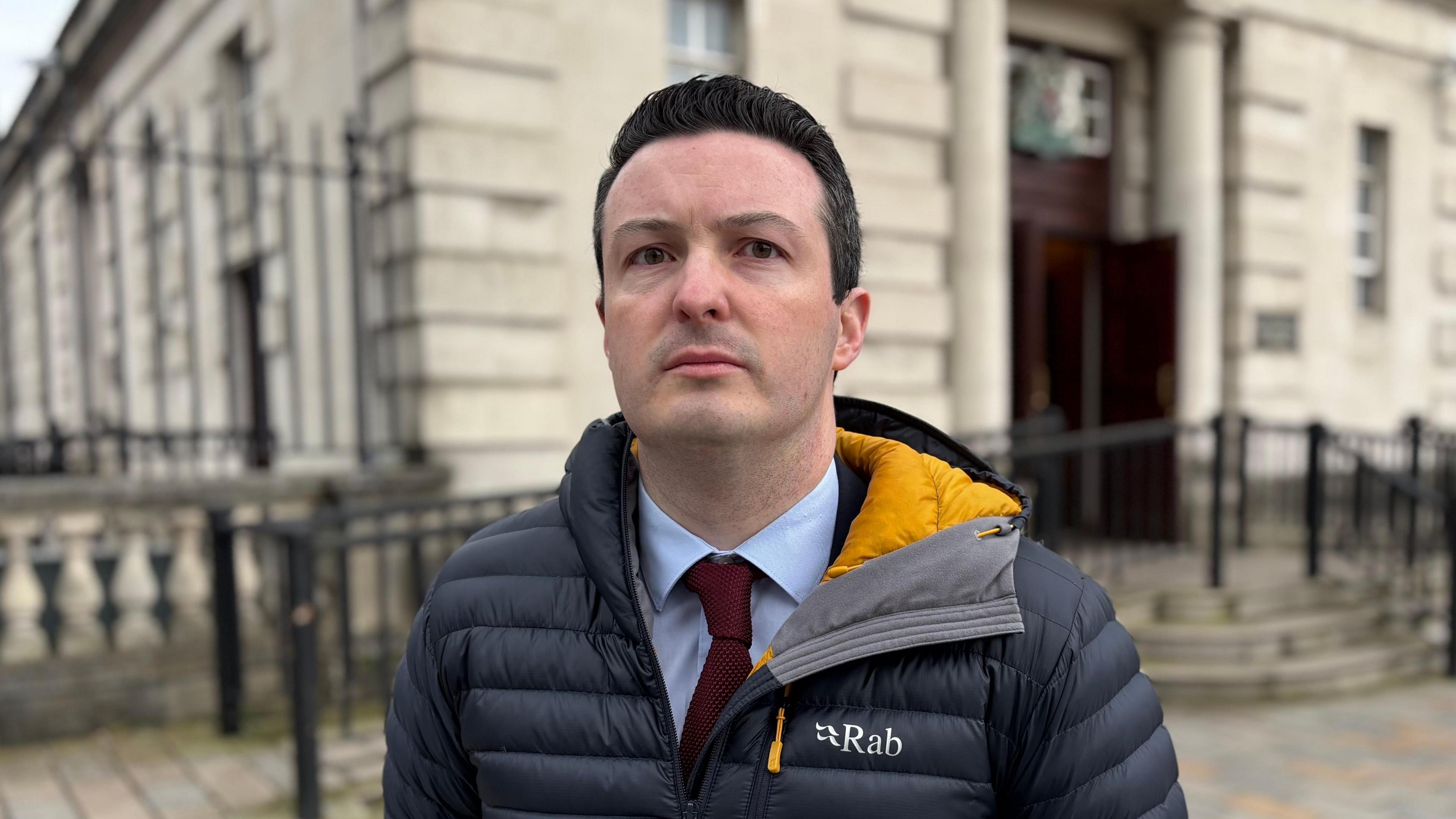 A man, with short black hair, wearing a blue shirt, burgundy tie and a navy coat. He is standing in front of a beige brick building.
