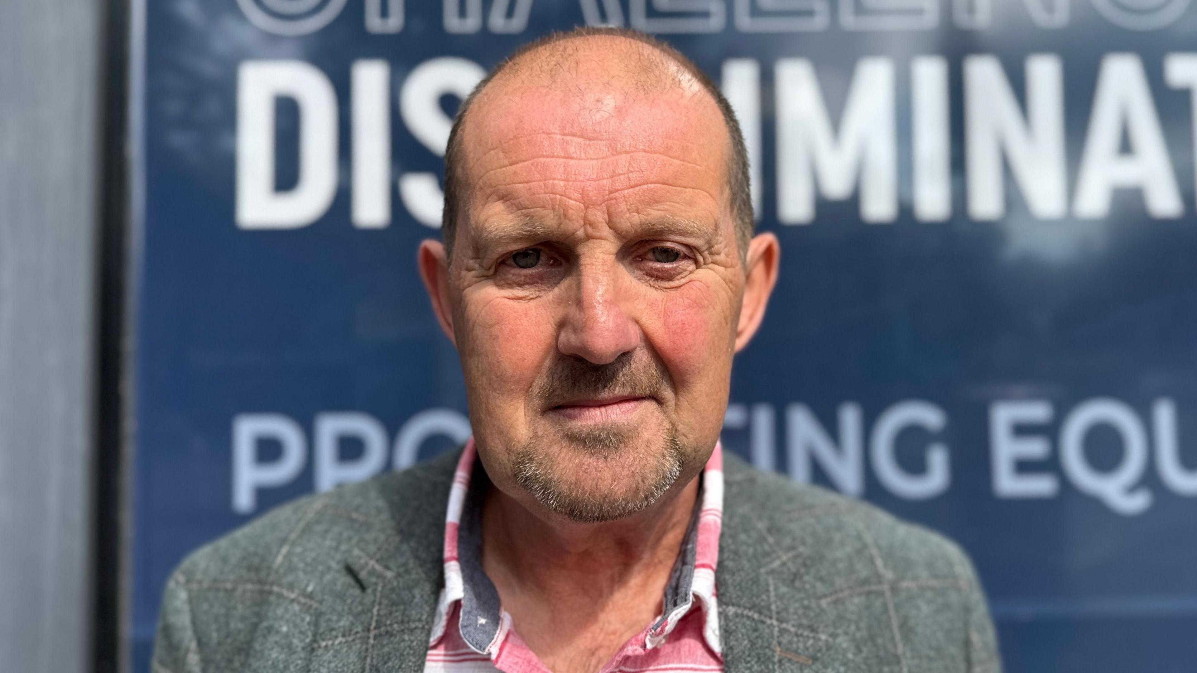 Cliff Donaldson is looking straight into the camera with a neutral expression on his face. He is an older man with a bald head, and is wearing a grey plaid blazer and a red and white flannel shirt underneath. He is standing outside the Equalty Commission building's blue signs.