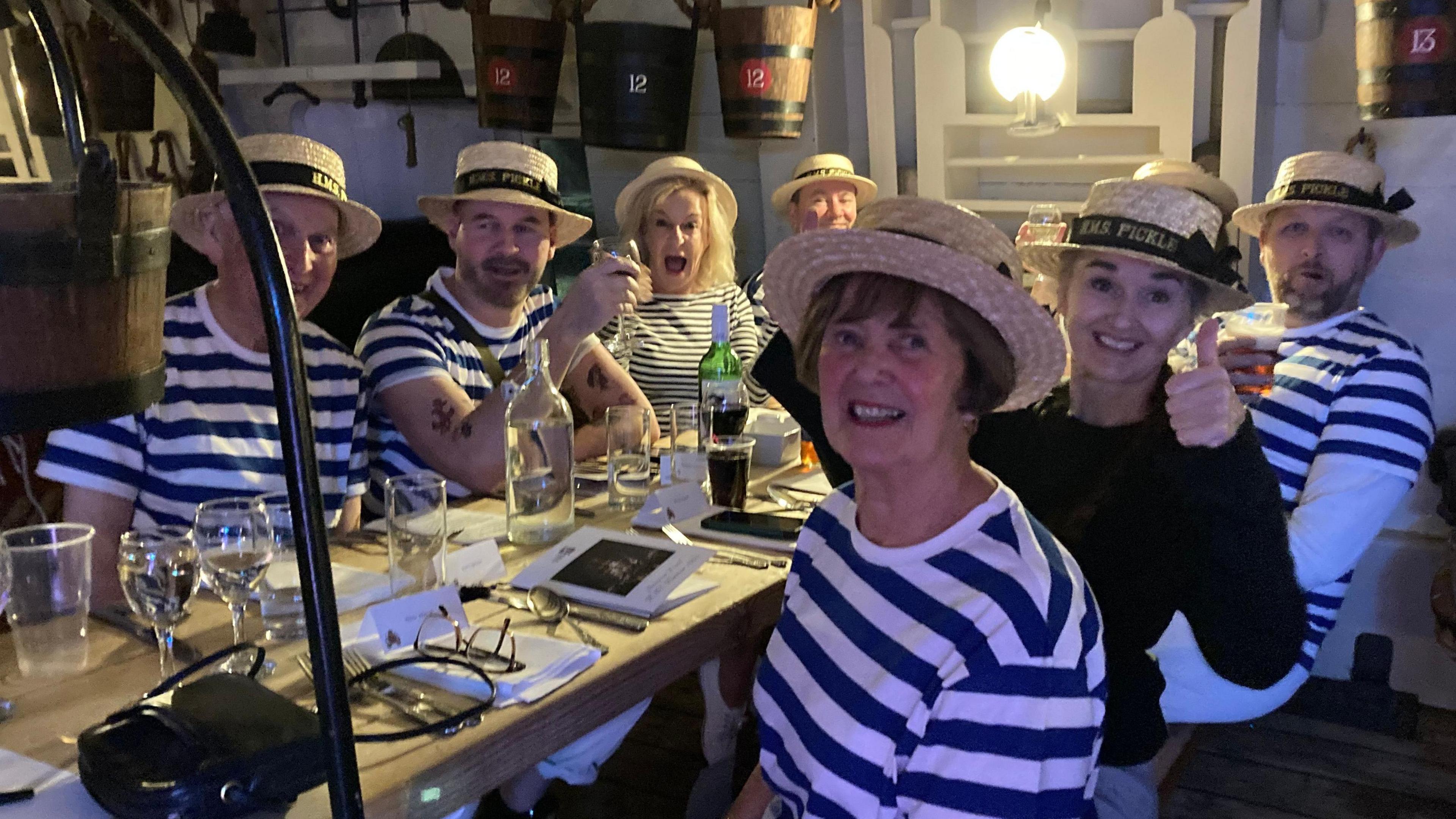 Half a dozen men and women, dressed in blue and white striped tops, red scarves, white trousers and wearing a hat, all sat around a long bench-style table, smiling at the camera