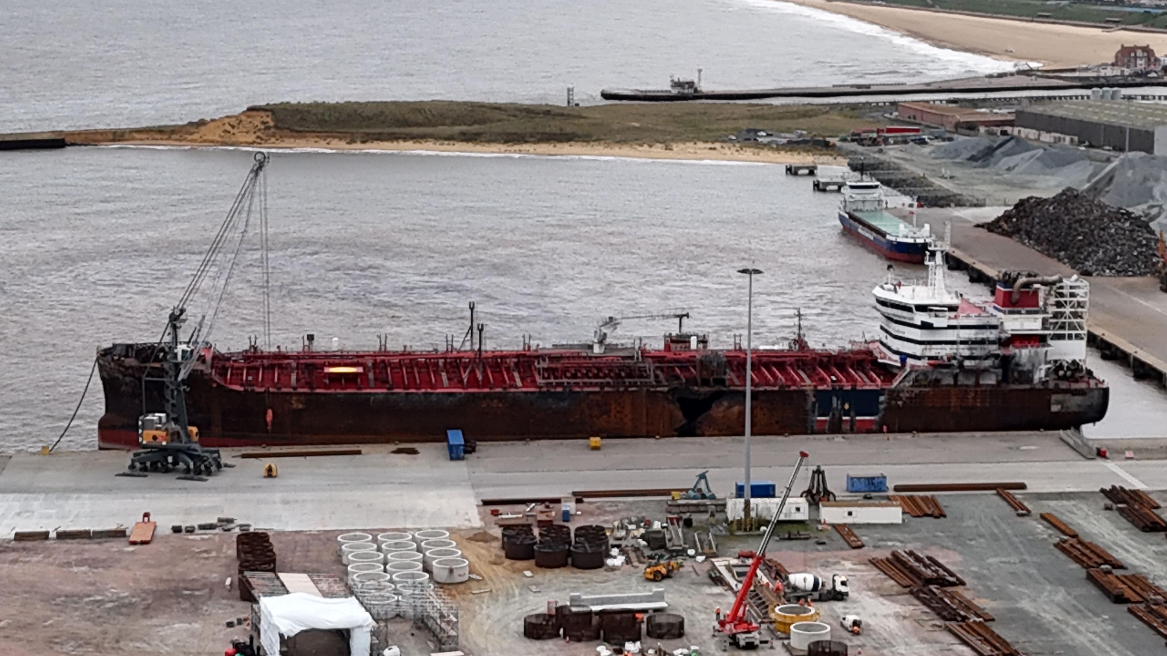 A drone image looks down on the red supertanker at anchor in a port, with equipment, machinery and materials on the land around it.
