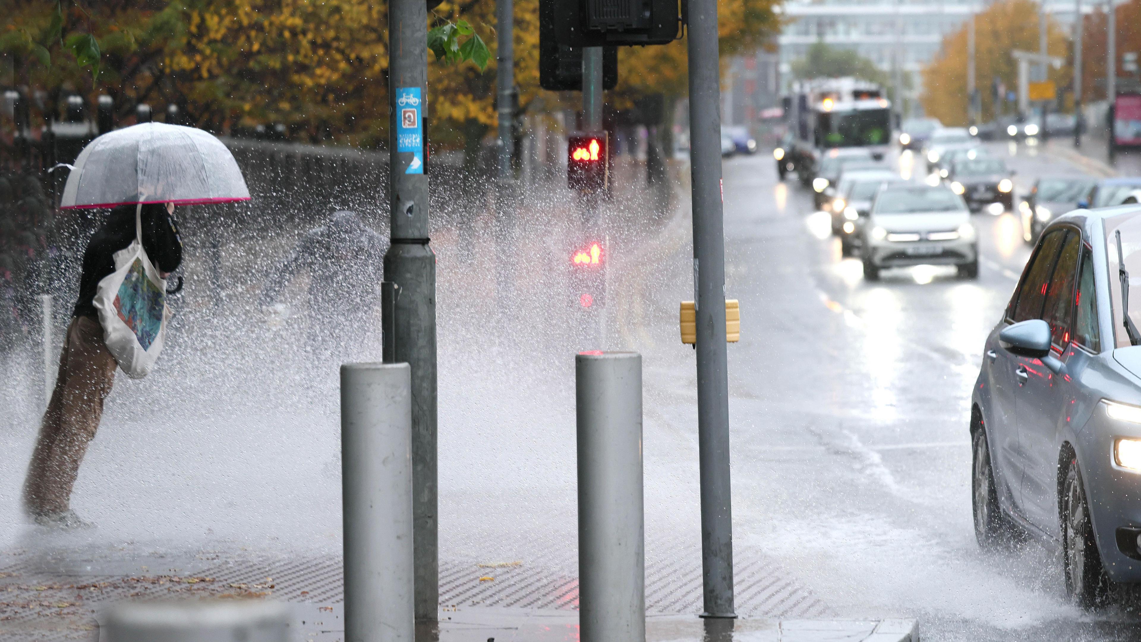 A pedestrian holding an umbrella stands at a set of traffic lights. They are leaning forwards as they are splashed by water from passing cars.