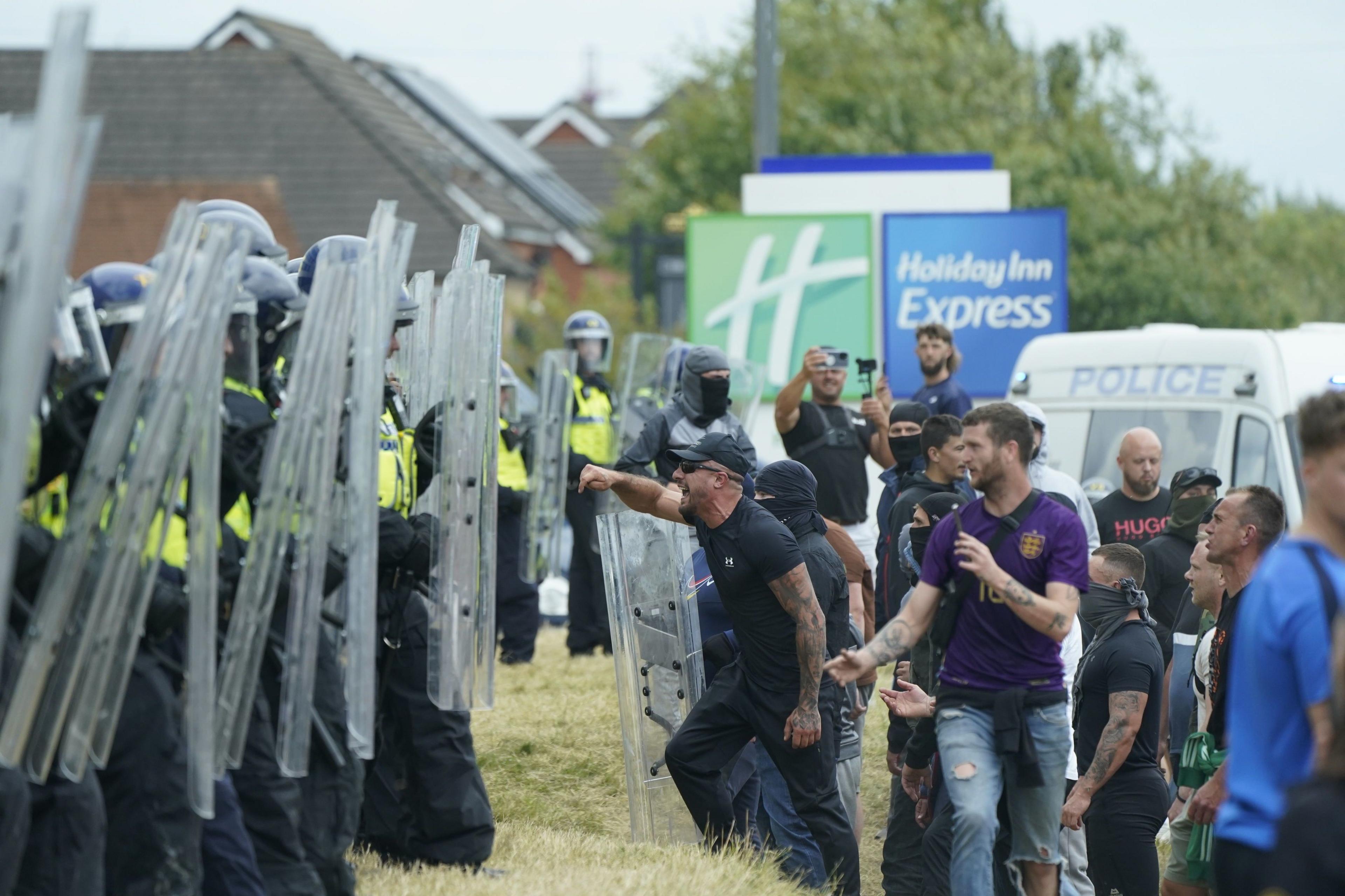 A line of riot police holding shields face off against demonstrators on a grass verge