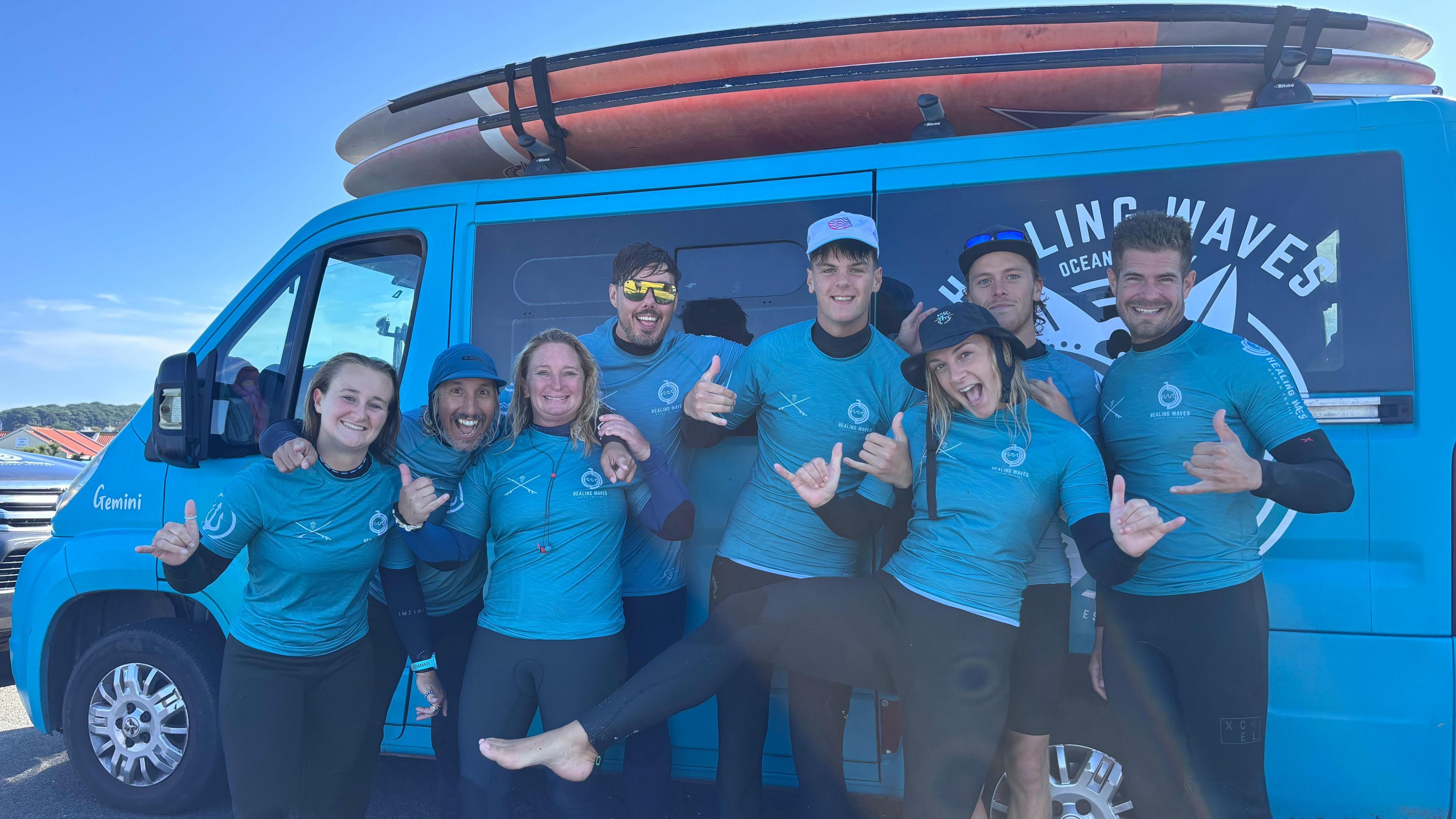 A group of smiling people stand in front of a van with an orange surf board on top. They are wearing pale blue tops and dark trousers. The words Healing waves can just be seen behind them on the van. 
