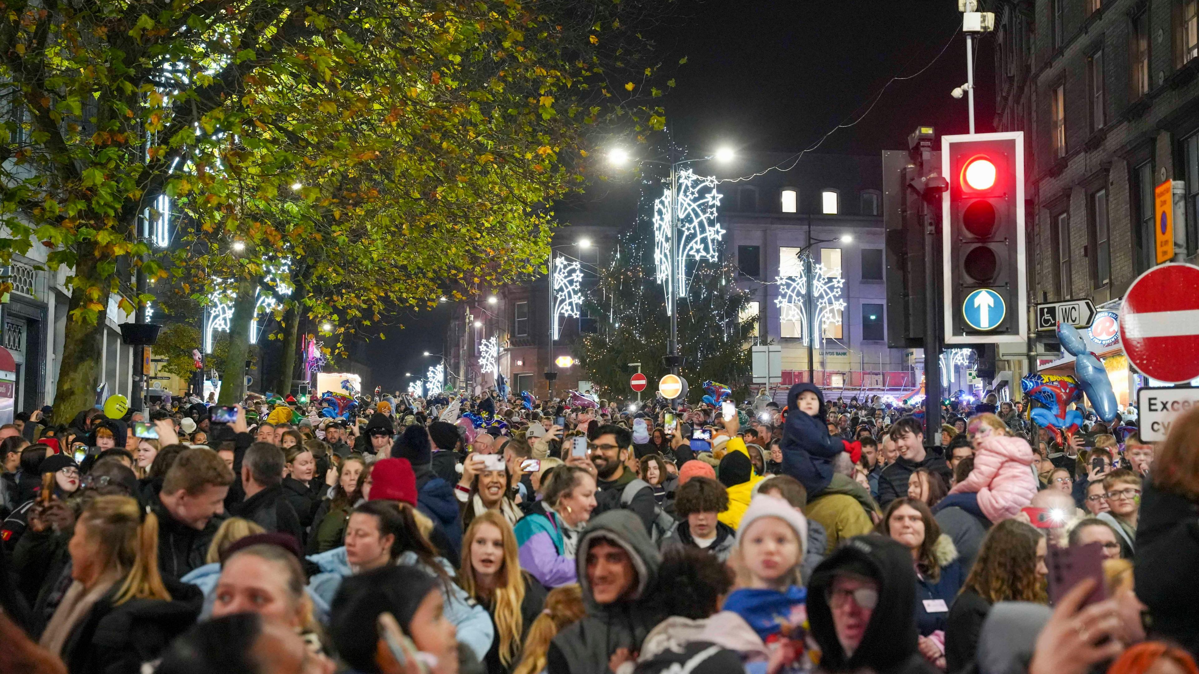 Crowds of people in Wolverhampton city centre, with road signs and a traffic light, along with trees in the background and Christmas lights lit up overhead.