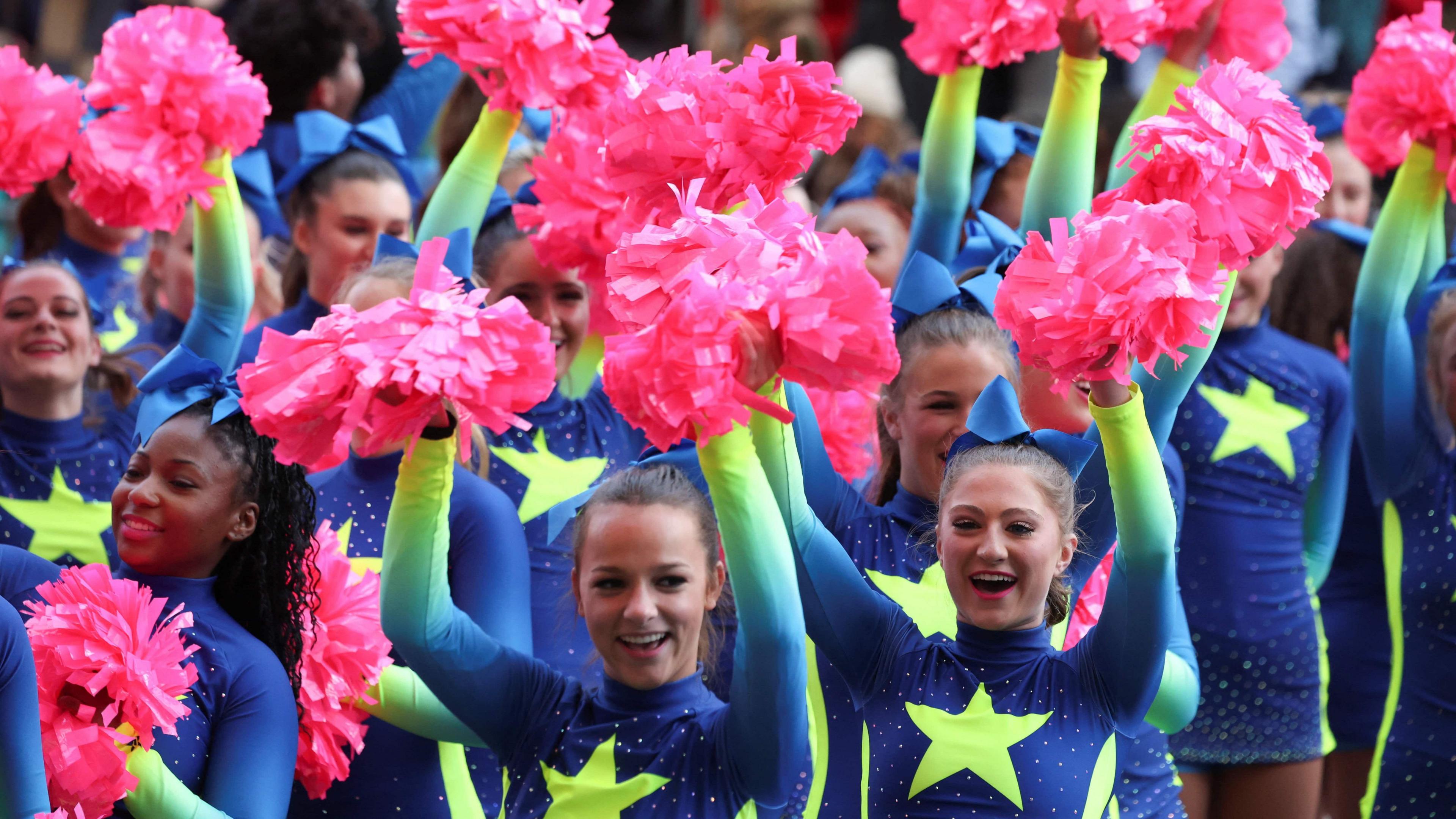 Performers wearing blue and green cheerleading outfits wave bright pink pompoms in the air. They are all smiling and cheering.
