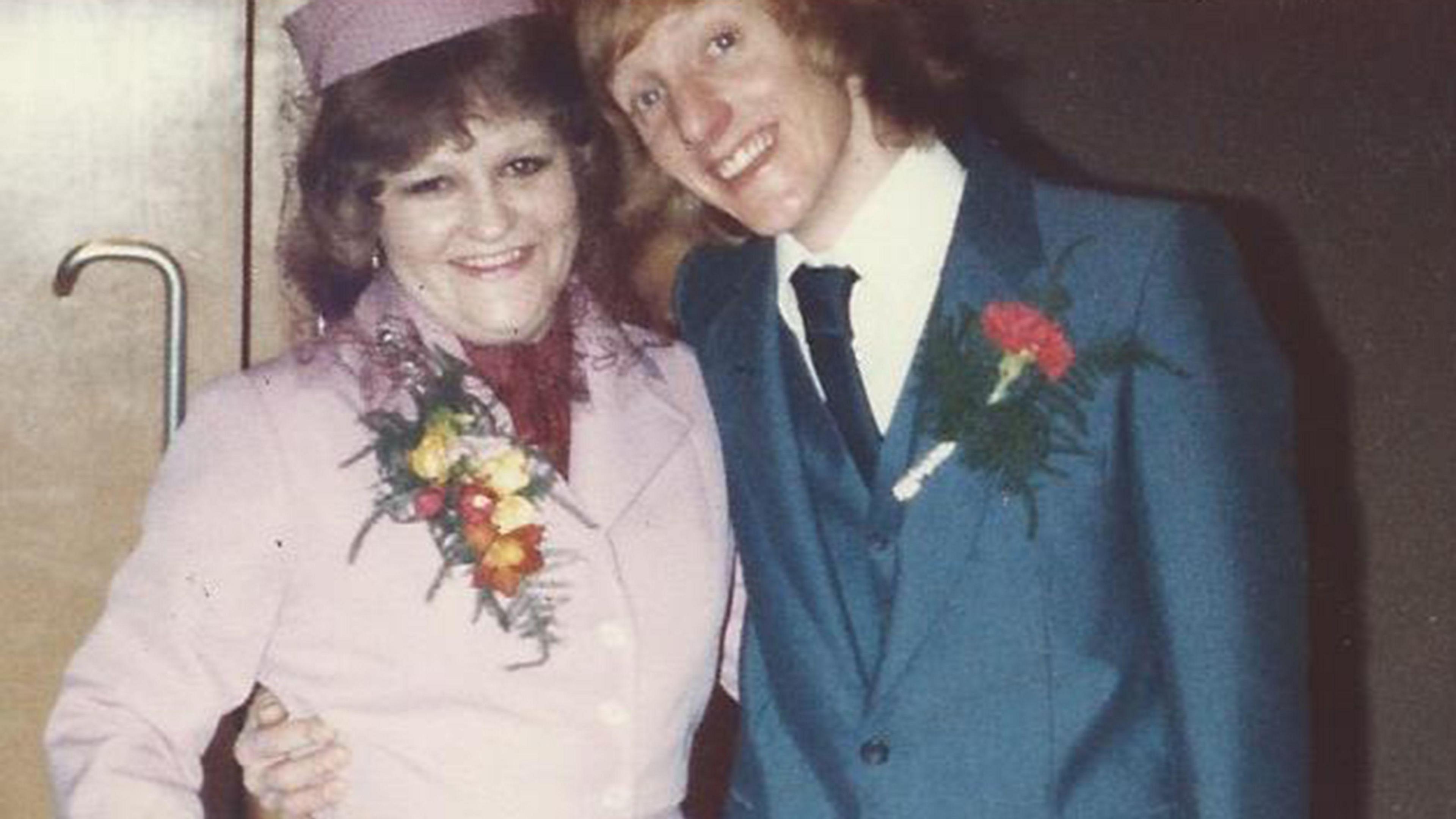 Patricia and Keith standing closely side by side against a plain interior background. Patricia is wearing a light pink, formal coat with a matching hat and a floral corsage pinned to the front. Keith on the right is dressed in a teal or blue formal suit with a white shirt and dark tie, and a floral boutonniere is pinned to the jacket