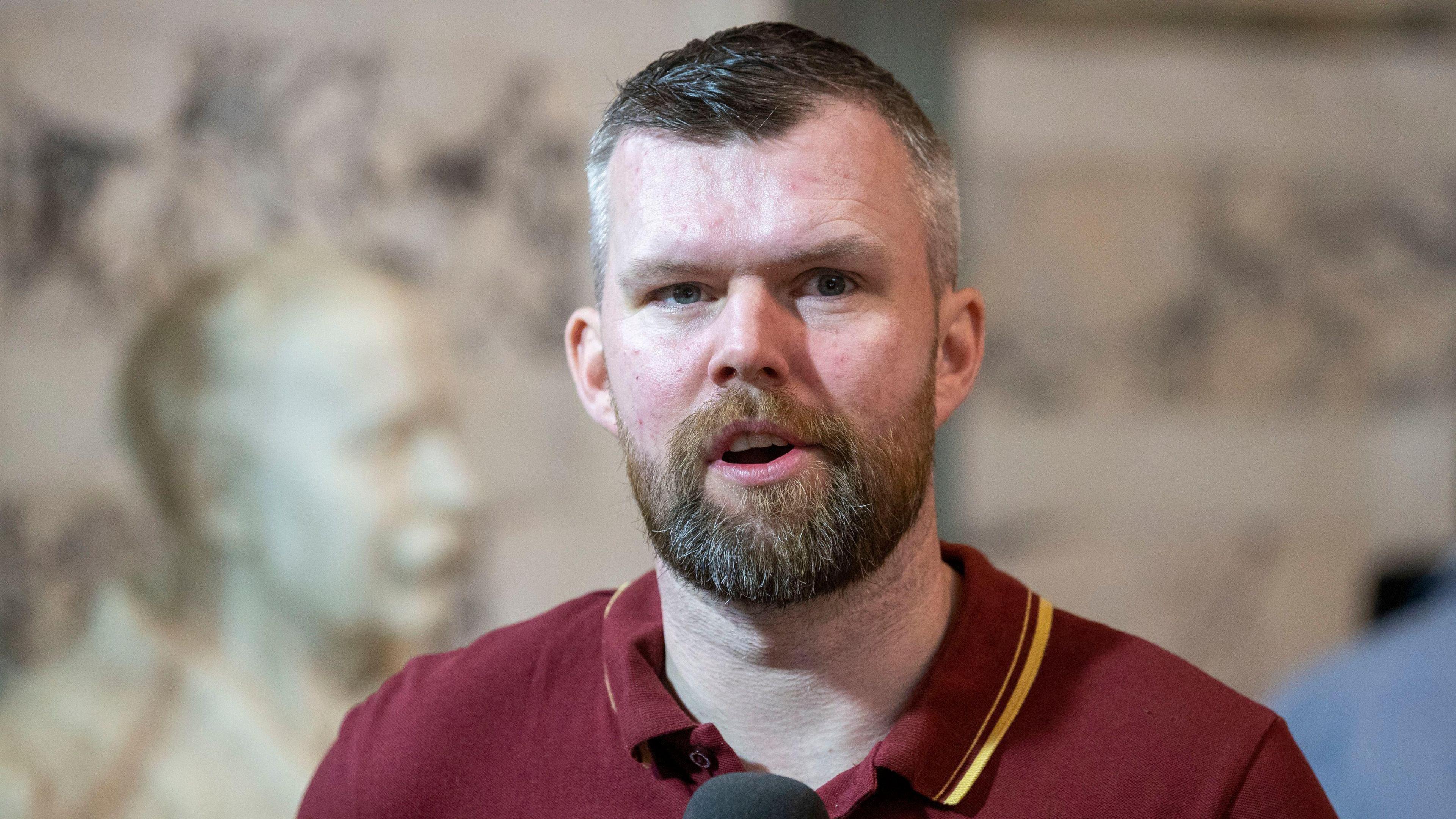 A man with short dark hair and beard is speaking into a mic wearing a red polo shirt.