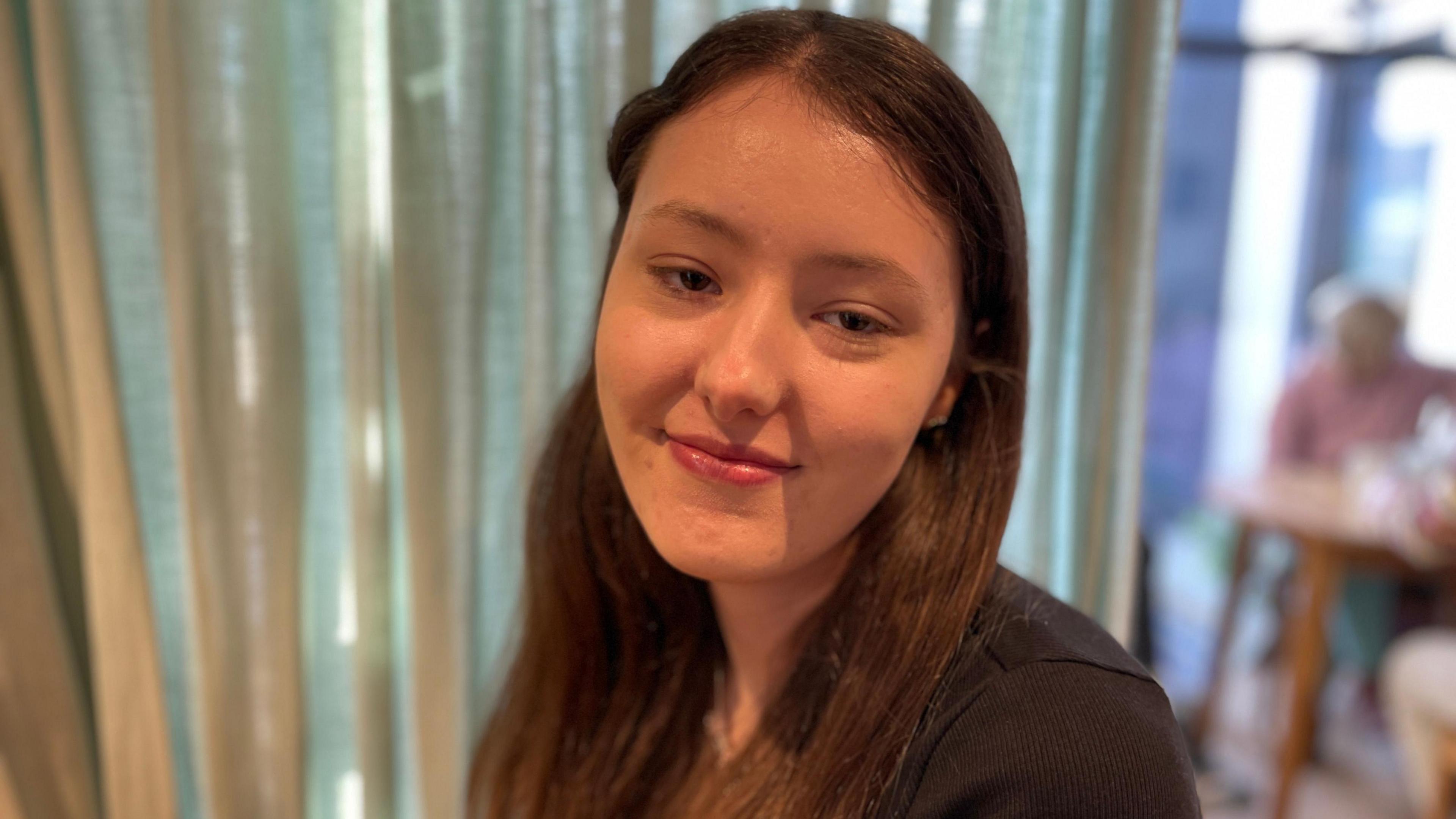 Lauren is a young woman with long brown hair which she is wearing down. Her hair is tucked behind her ears and she's wearing a black top and looking at the camera smiling. Behind her is a pale green curtain.