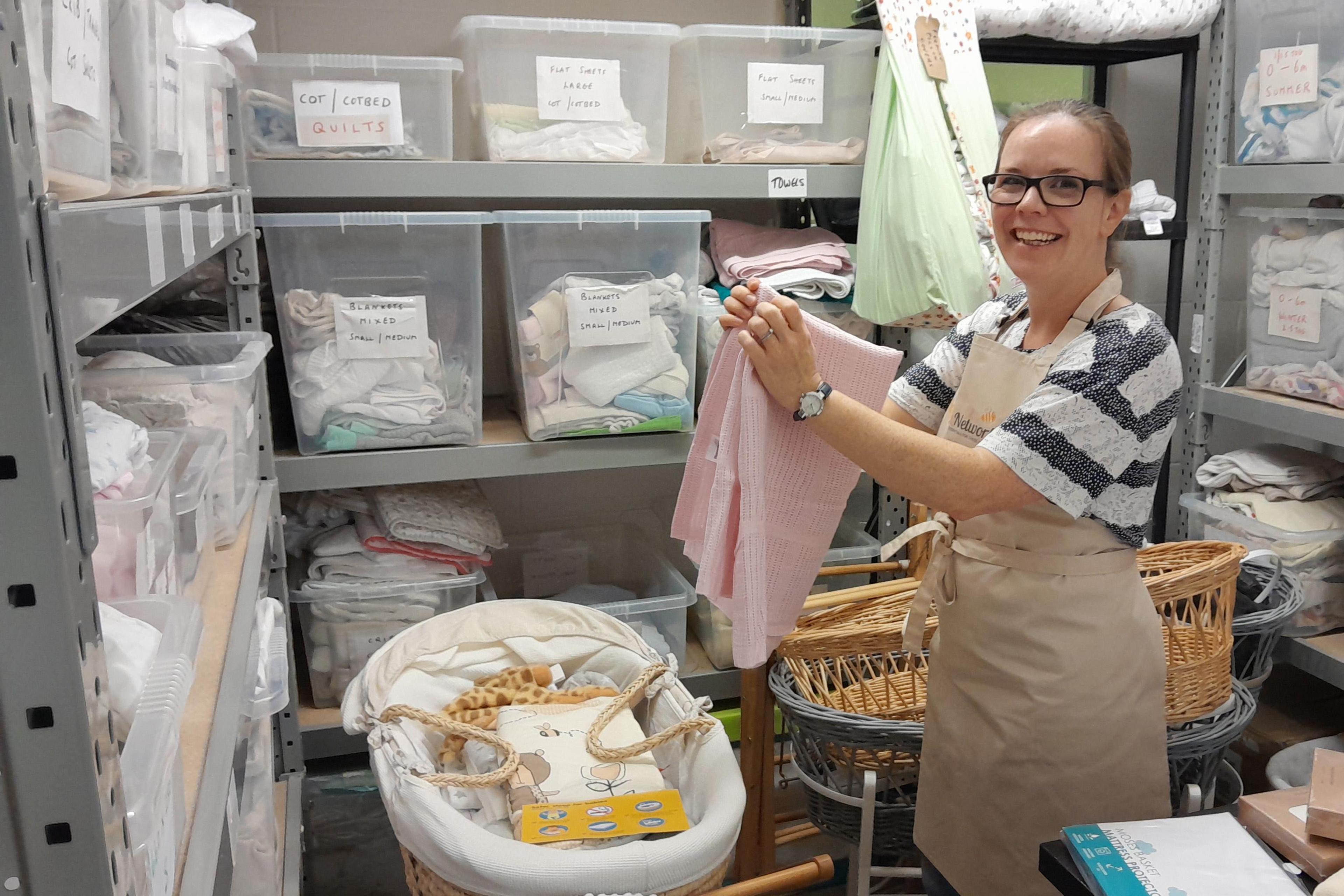 A woman is in the middle of a storage room folding a pink blanket. She is surrounded by three portable baby cots and shelves packed with labelled clear plastic boxes containing folded baby items.