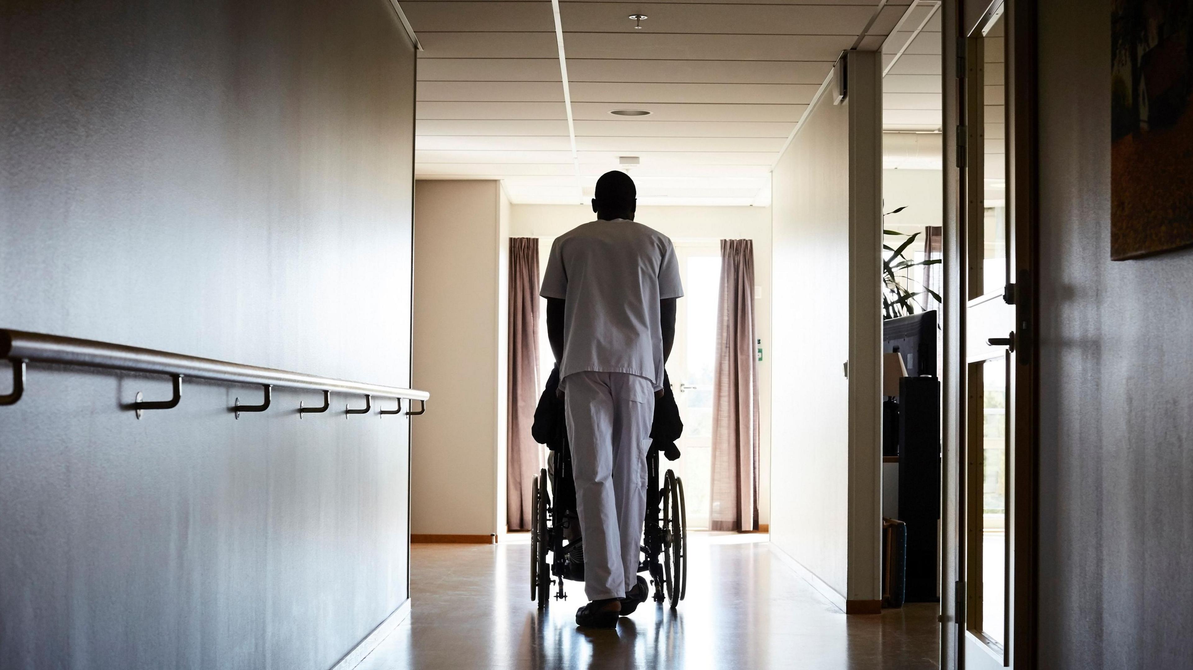 We see the back of a person wearing white pushing a wheelchair along a corridor