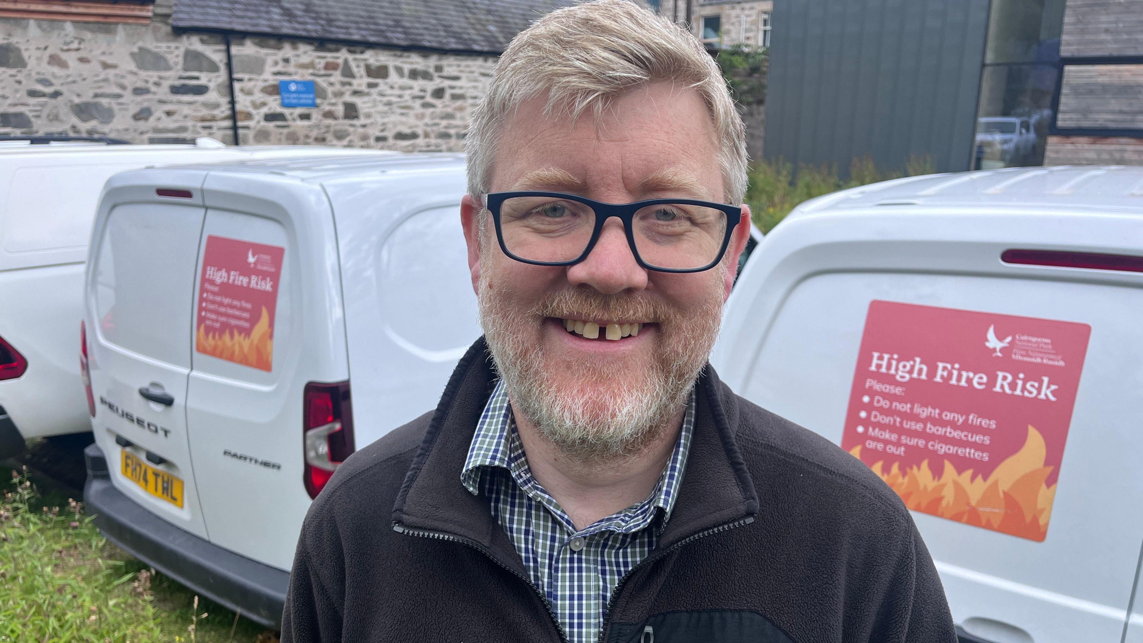 A mark with dark glasses and a beard, wearing a fleece and checked shirt, stands in front of two white vans displaying warnings of a high fire risk.