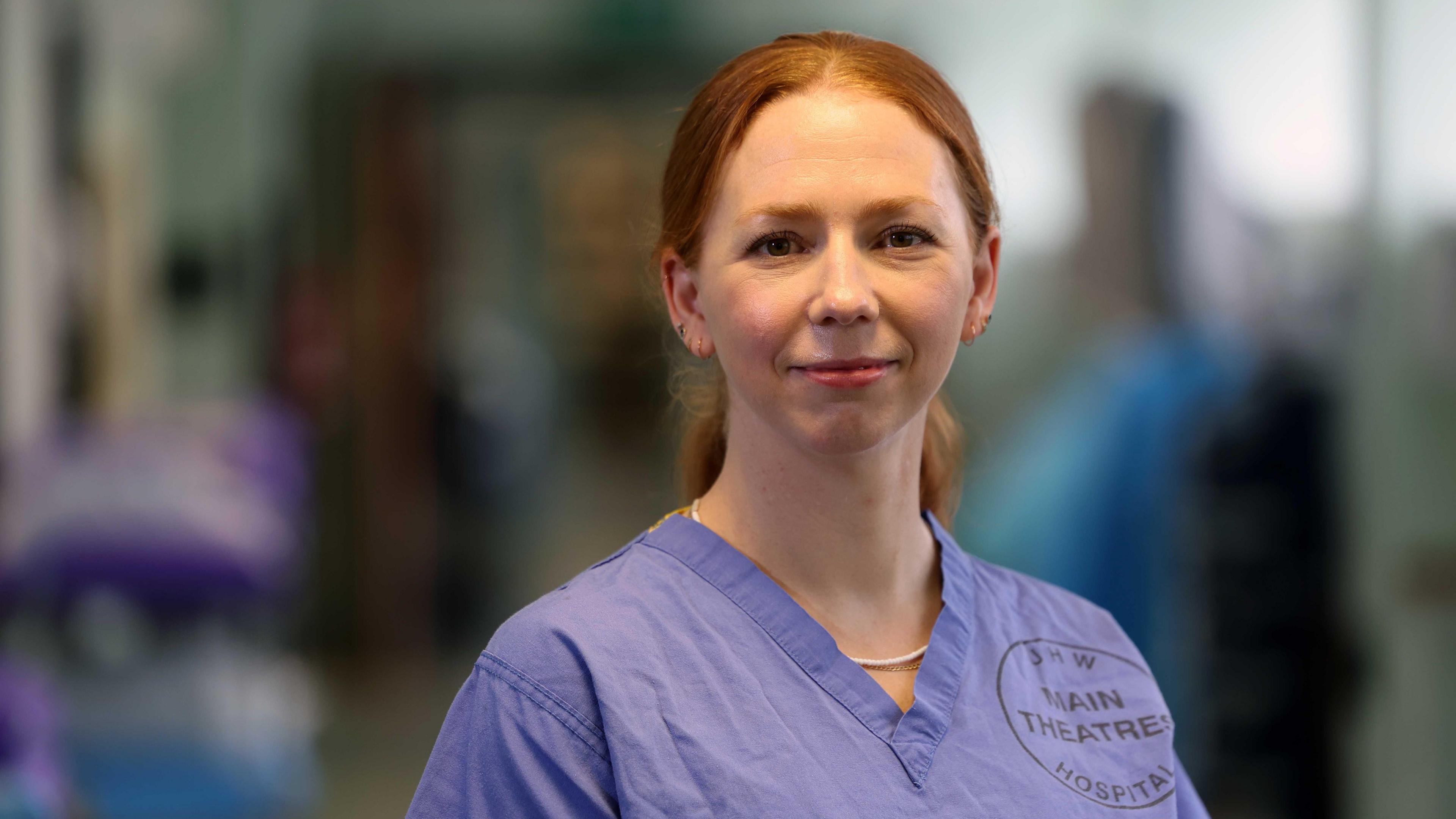 Jody is wearing blue theatre scrubs and there is a blurred hospital corridor in the background.