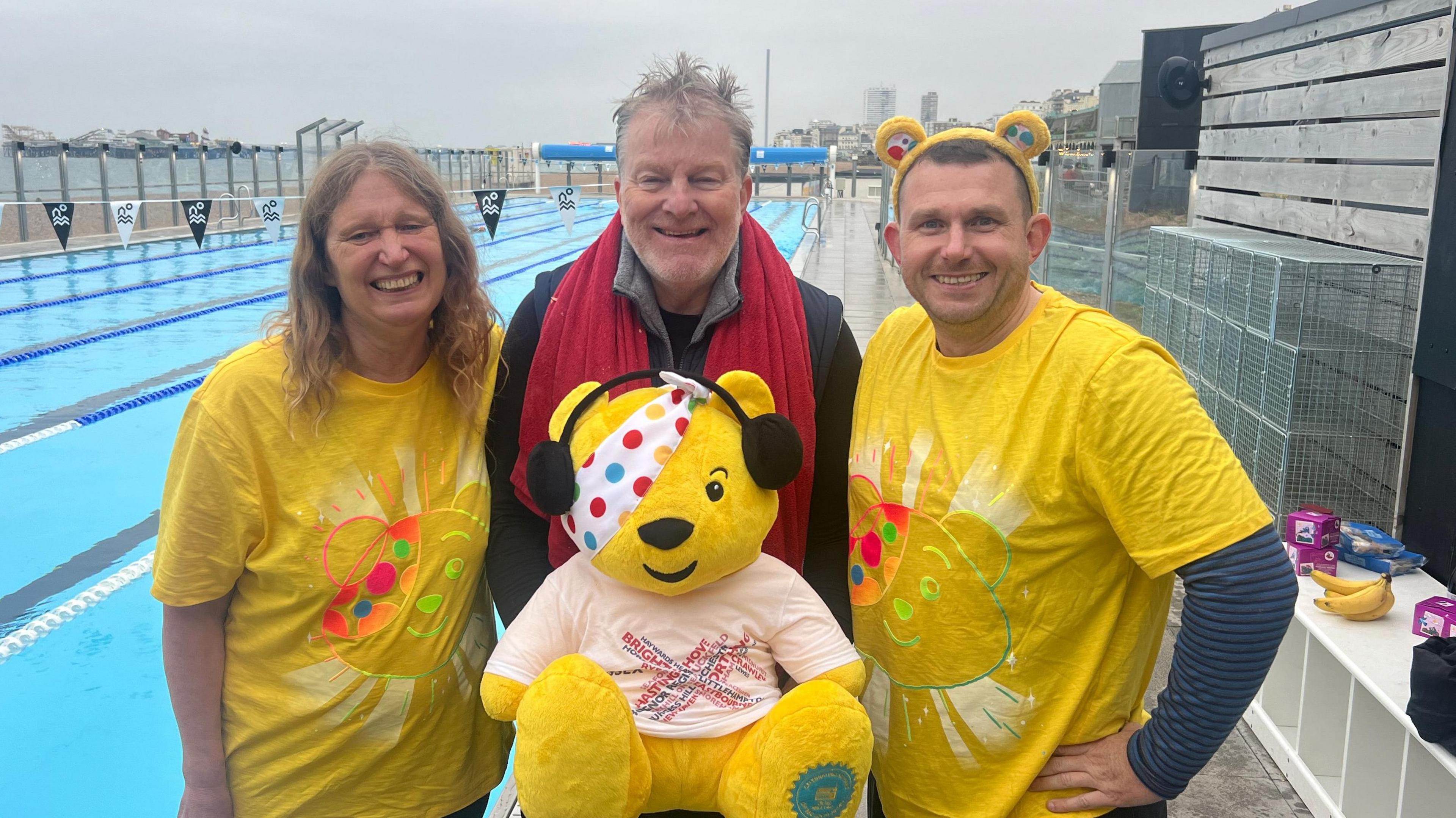 A man and a woman wearing yellow Pudsey shirts. The man has Pudsey ears on too. They are either side of a man with a red towel round his neck holding a Pudsey teddy bear which has a BBC Radio Sussex shirt on.