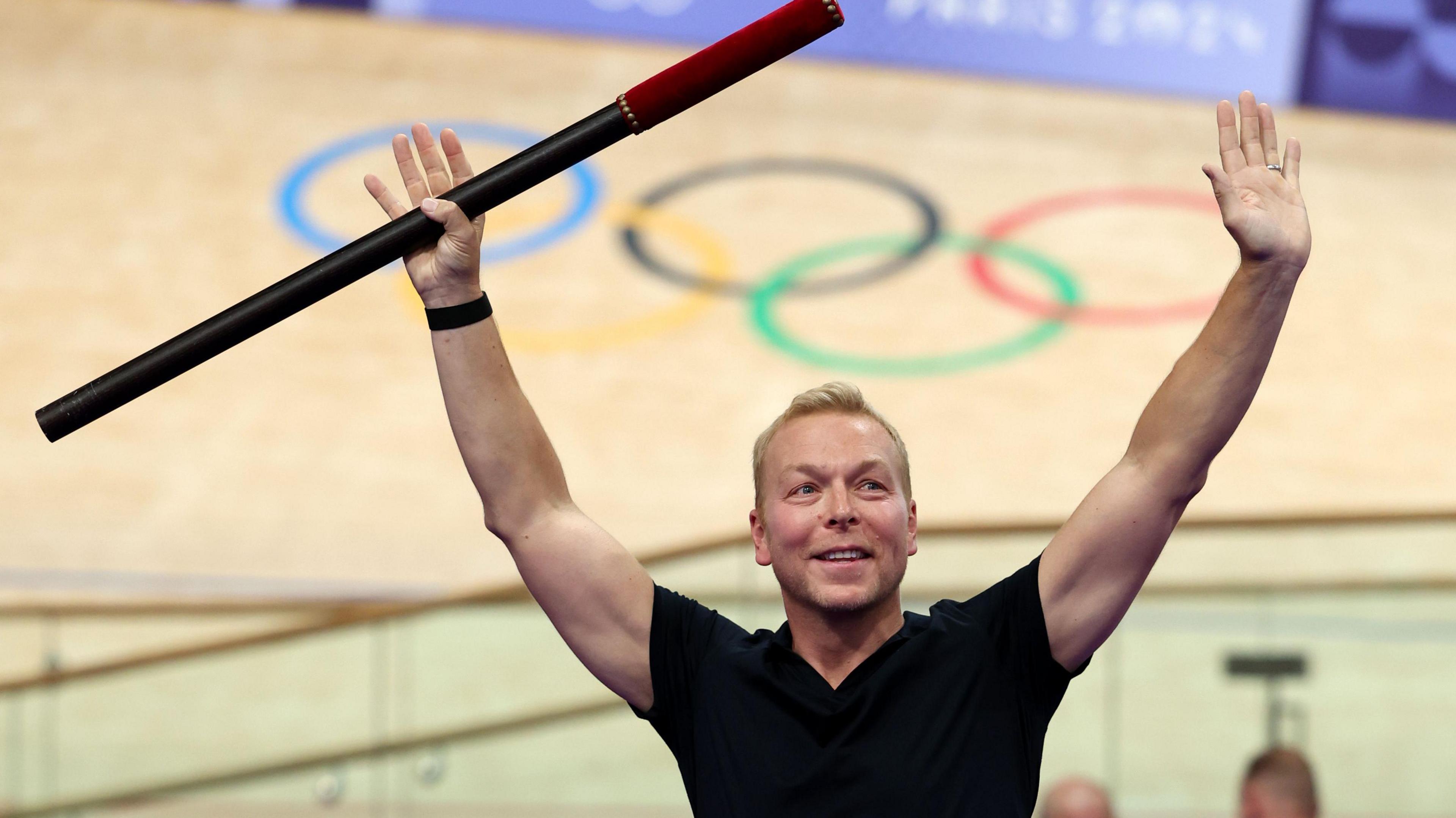 Chris Hoy holds a baton in the air during a ceremony at a velodrome in Paris. Behind him is the velodrome with the five Olympic rings