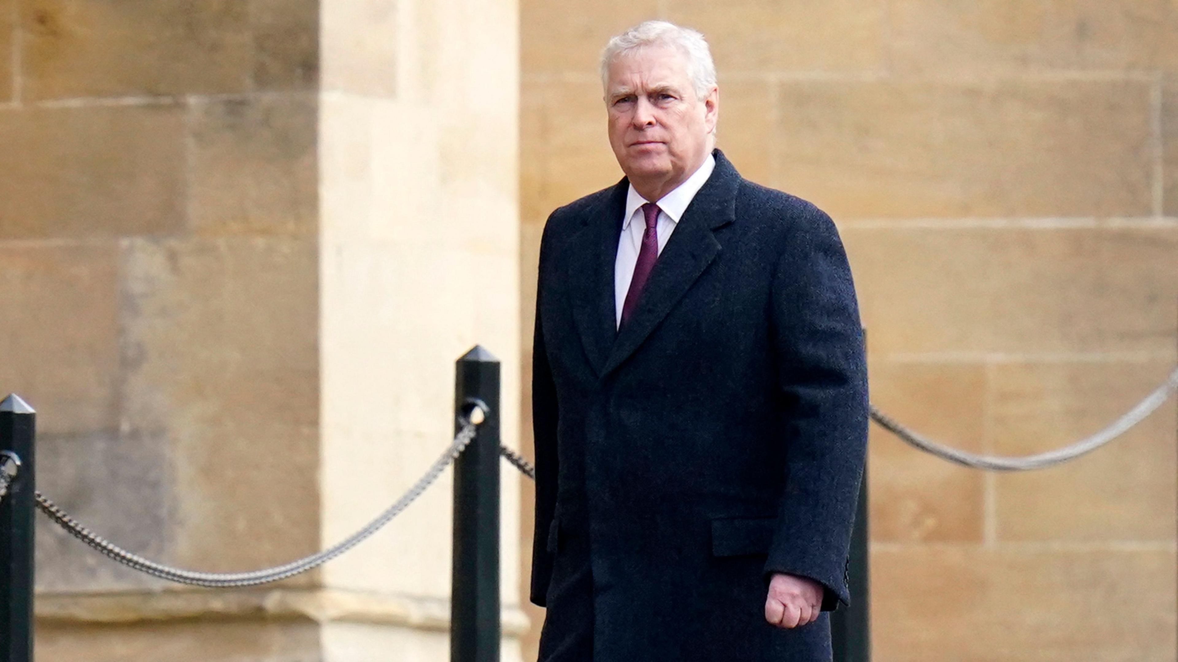 Prince Andrew looks towards the camera while walking in a suit and tie against a stone building backdrop, at Windsor Castle in February.