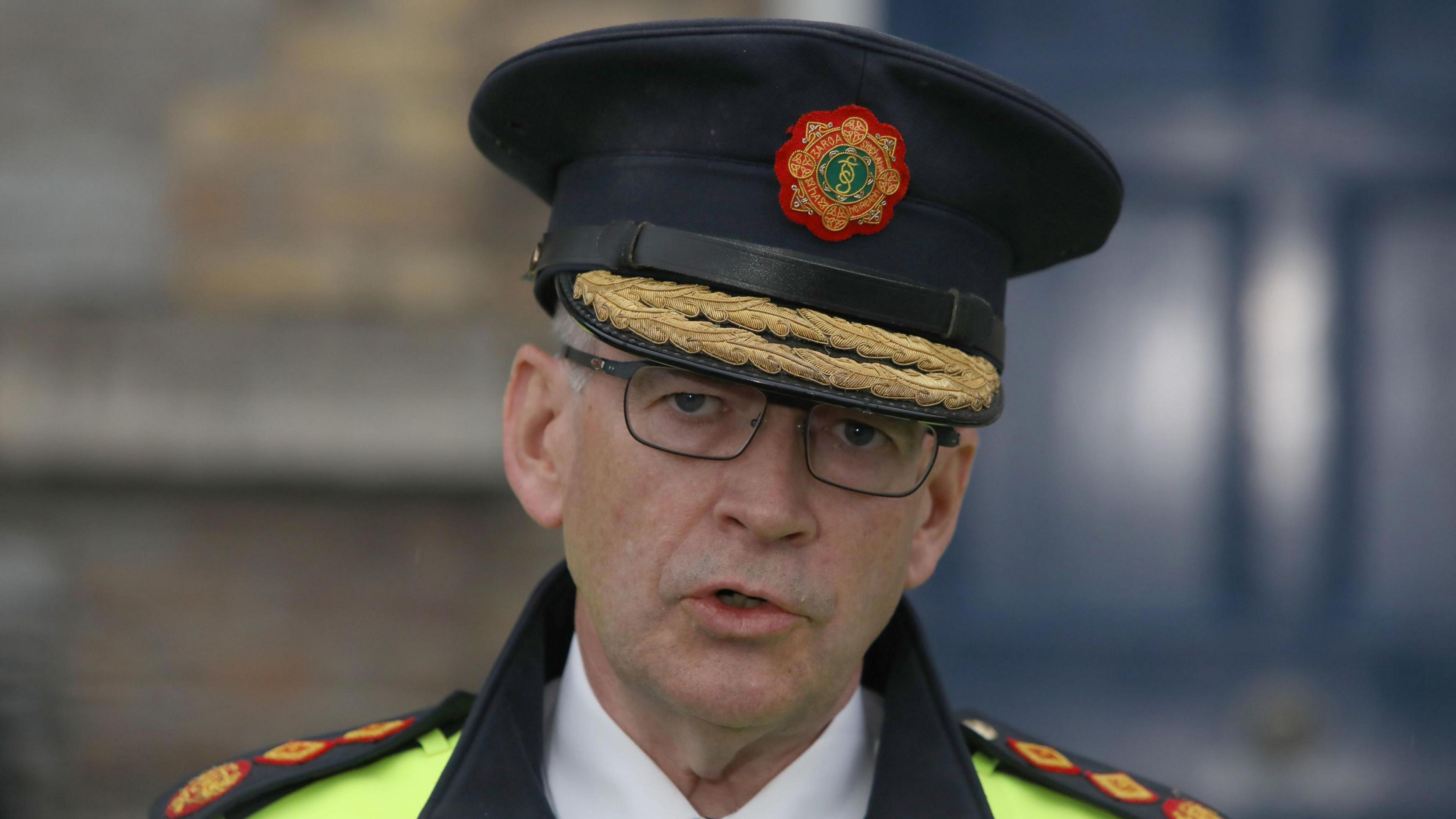 A close-up image of Garda Commissioner Drew Harris. He is an older man who is wearing a black, flat top hat with the garda logo in the centre and two gold ropes on the brim. He is mid-speech and is wearing a pair of grey metal glasses. He is also wearing a reflective police jacket and a white collared shirt.
