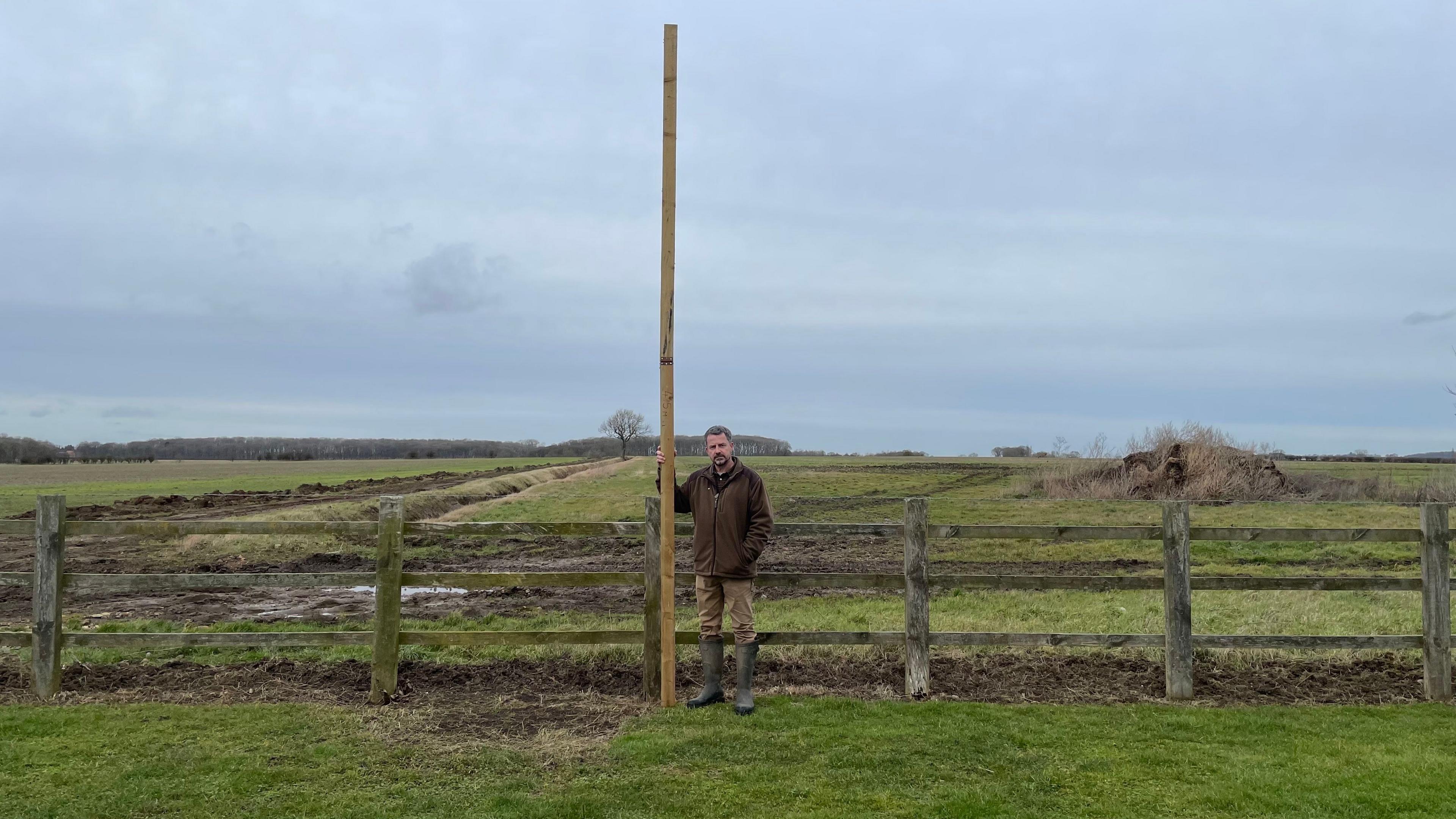 A man in a brown coat and light brown trousers, wearing wellies, is standing next to a tall post on farm land.