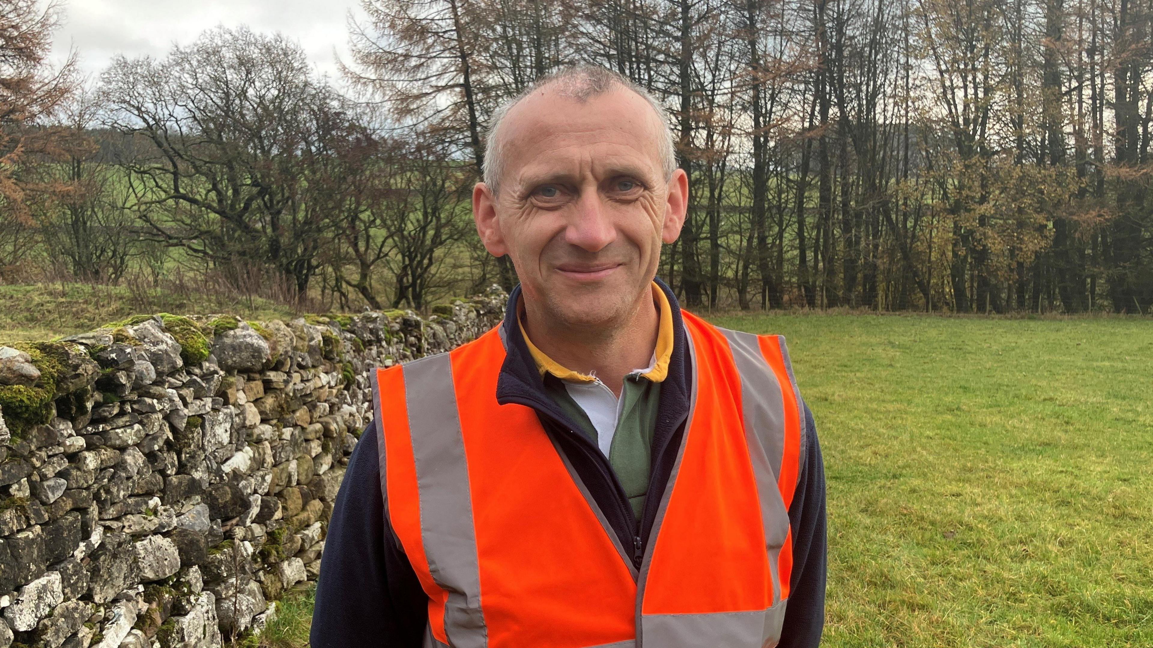 Ian Hutchinson is standing beside a dry stone wall in a field. He is smiling towards the camera wearing an orange hi-vis vest over a black fleece. In the background trees without autumn leaves can be seen against a background of the Cumbrian fells. 
