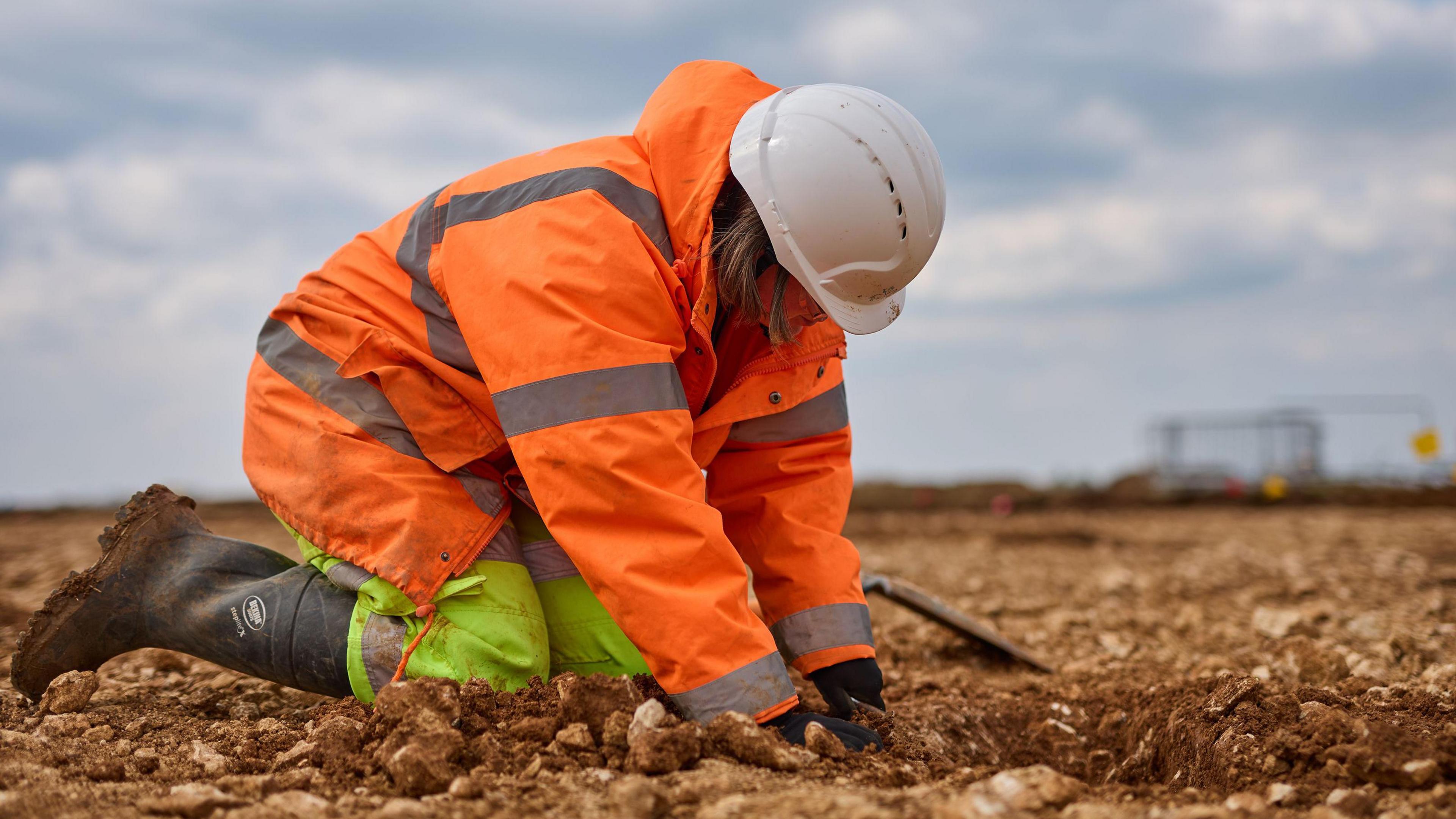 Worker digging up ground, they are wearing orange overalls and a white hard hat