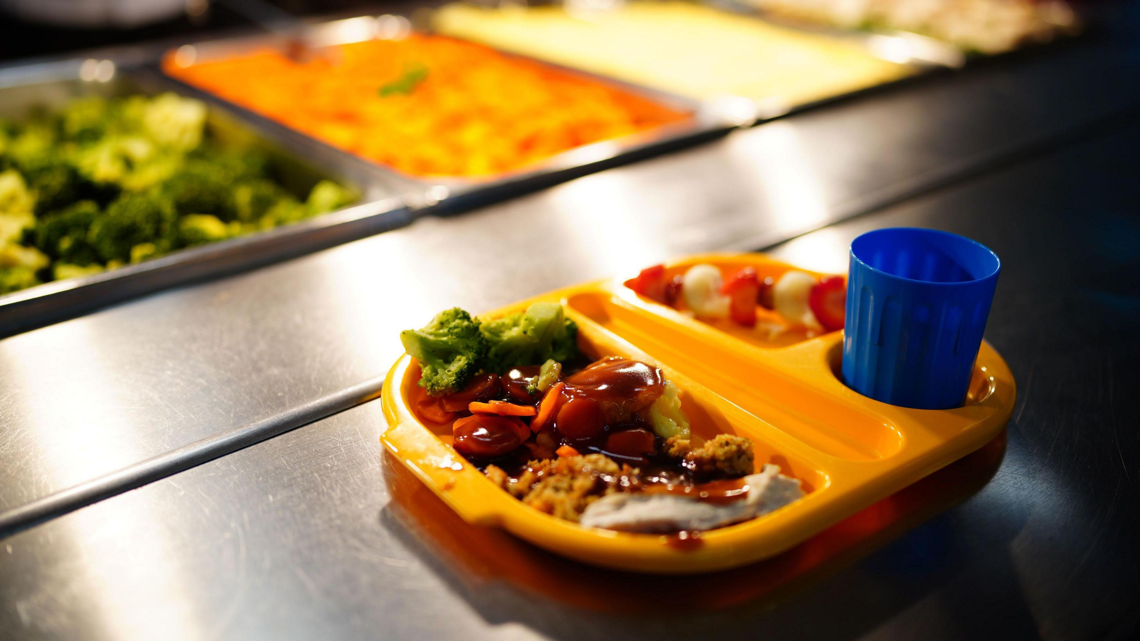 An image of a school dinner in a compartmentalised yellow tray with a drink in a blue plastic cup. The meal appears to be meat and gravy with broccoli and some fruit on a stick for dessert.