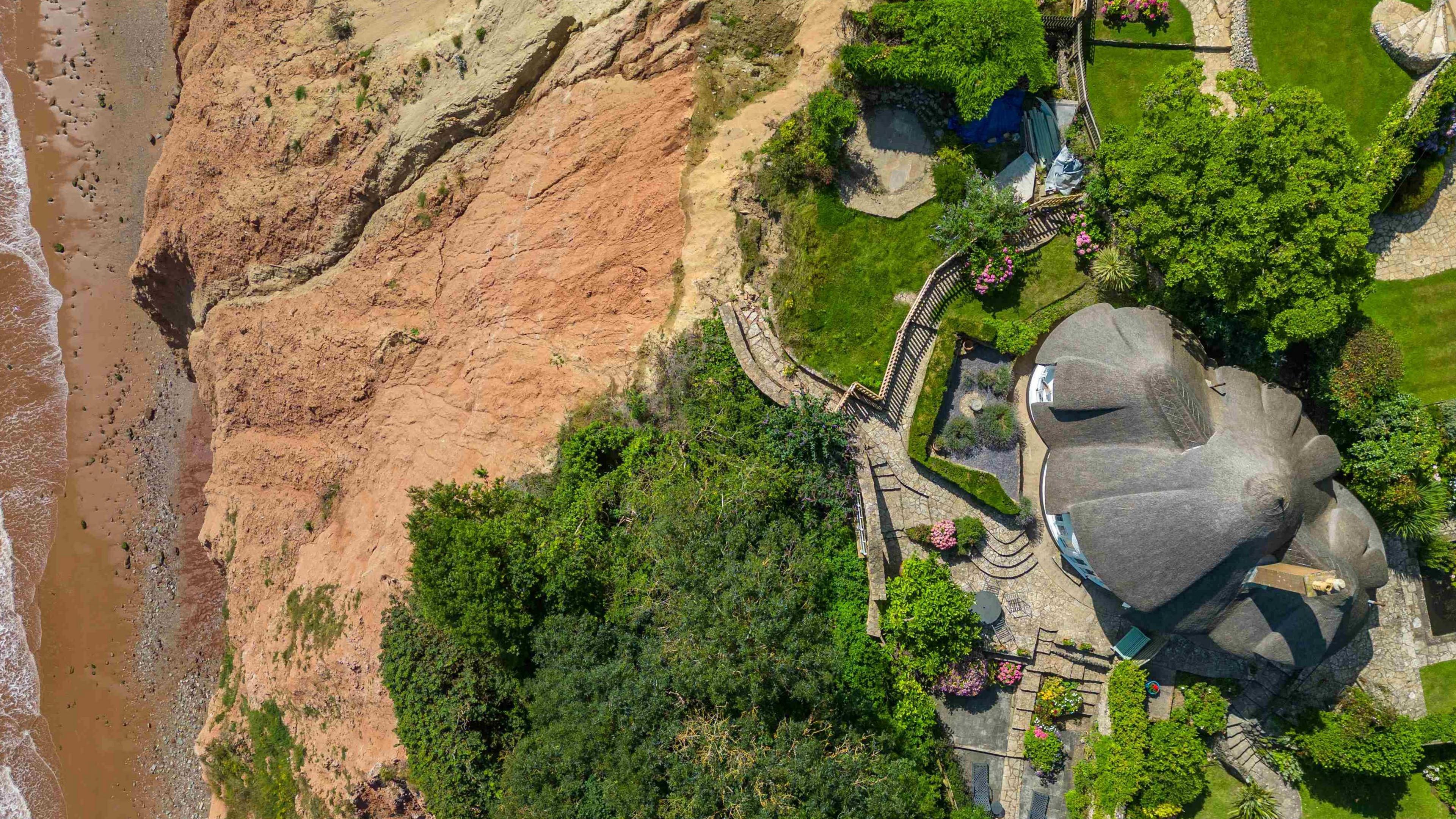 A house sits close to the edge above the sea with more houses further inland and nearby hedges and trees.