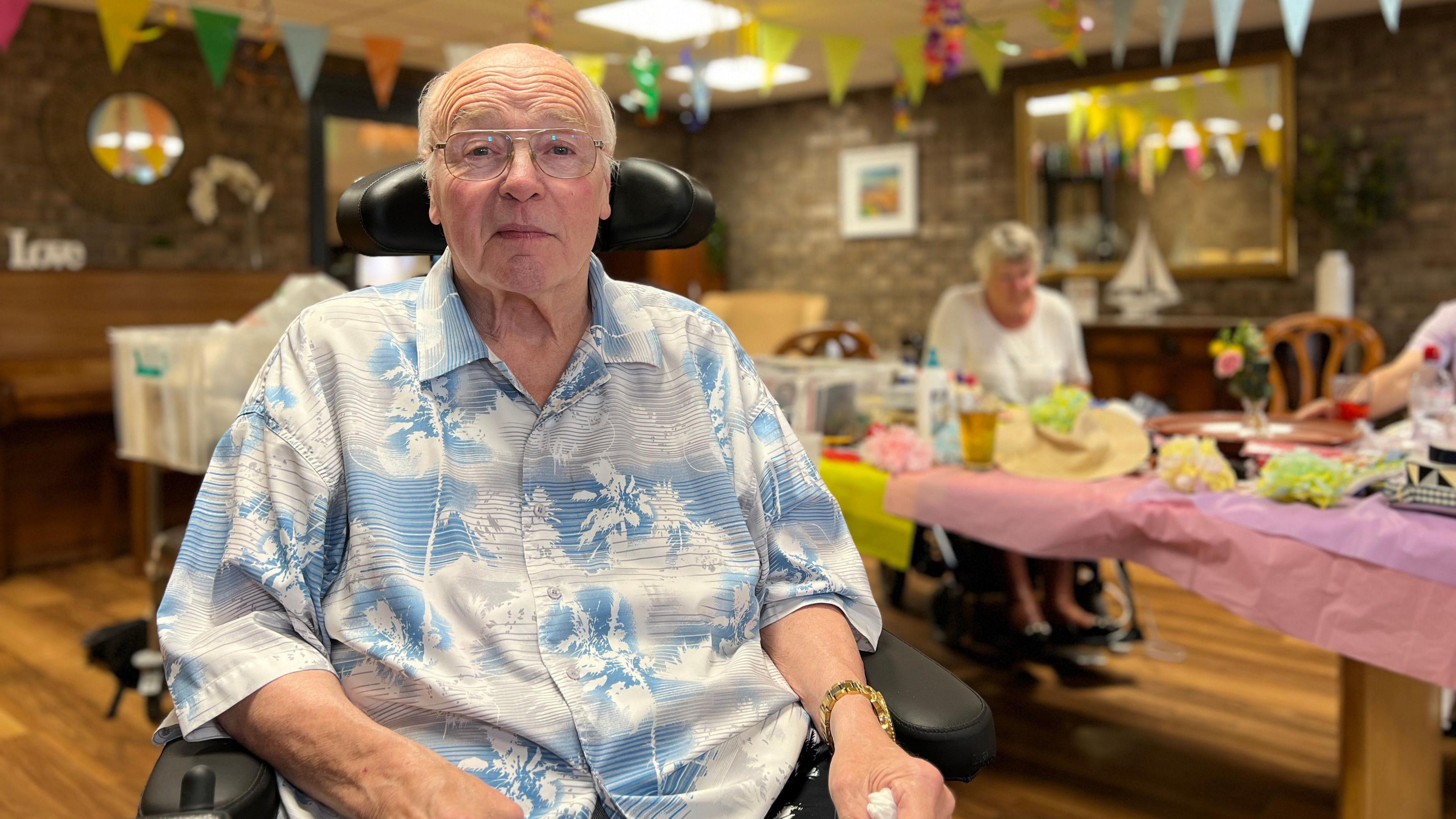 Stanley looks at the camera in his wheelchair as people work on the decorations behind him