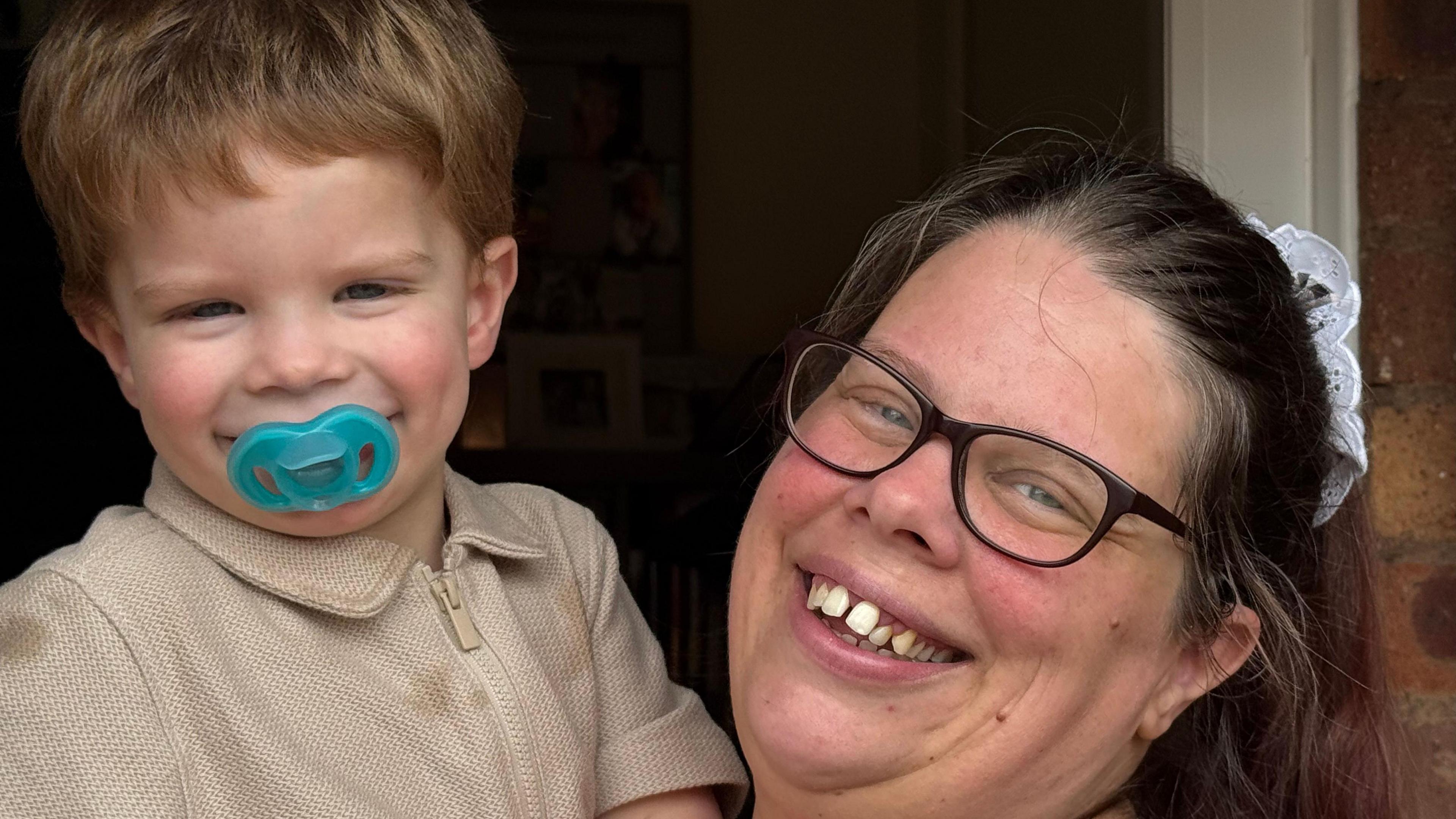 Stacey Harris holding her two-year-old son and smiling. She shows her teeth and wears black-rimmed glasses leaning her head against the side of a door. Her son has a turquoise dummy in his mouth and wears a beige top.