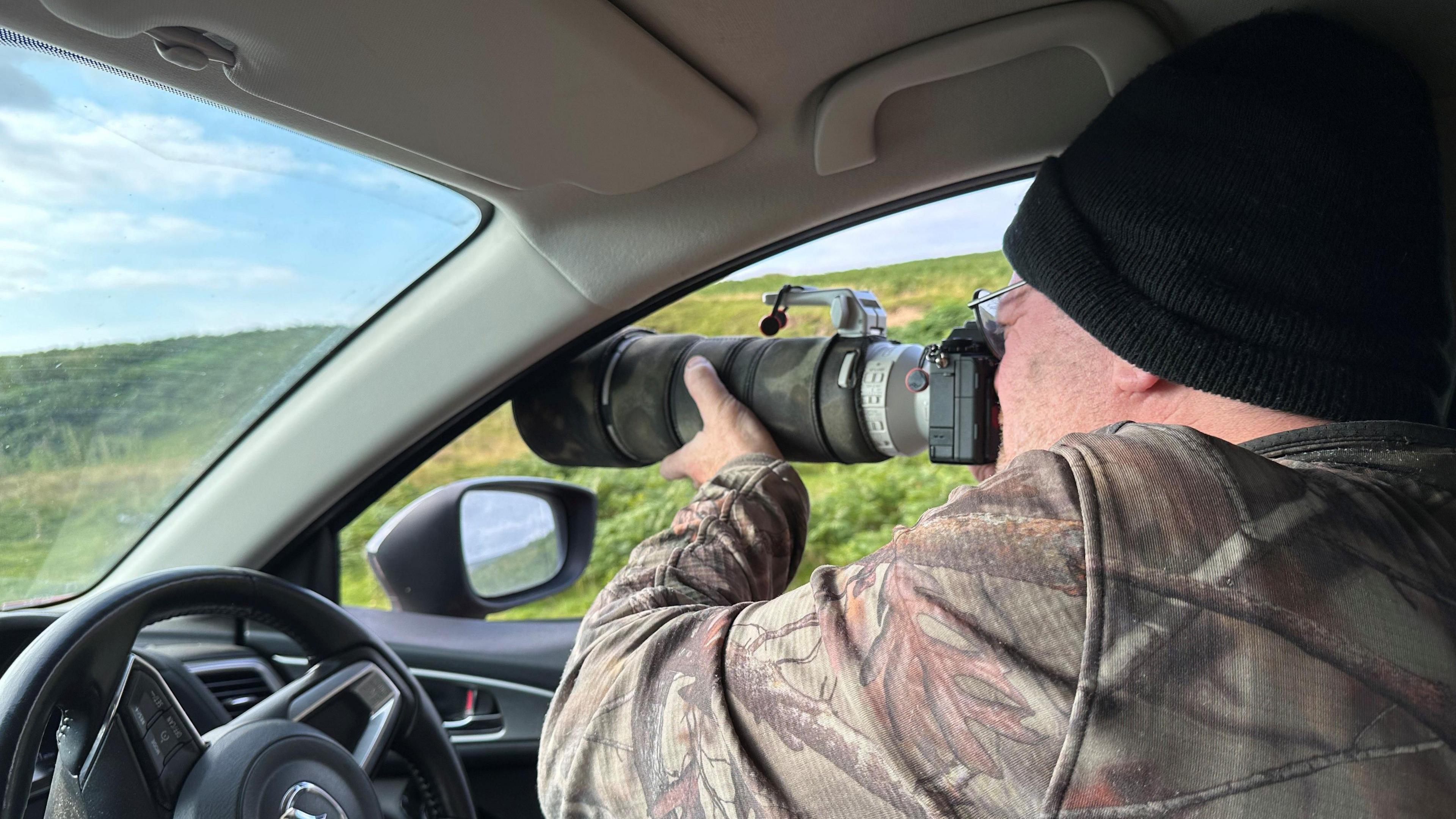 A man in a camouflage jacket points a camera out of a car window.