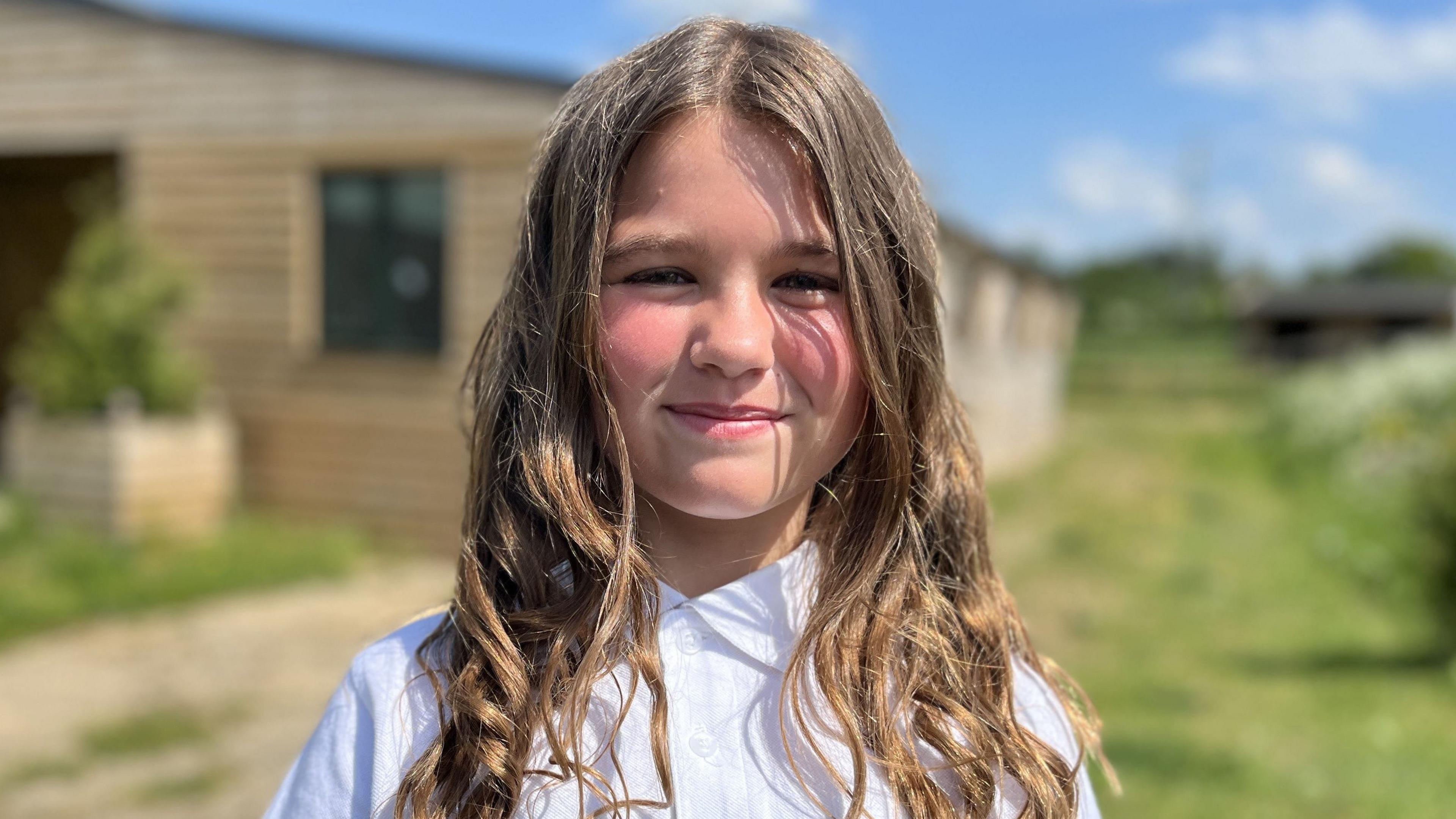 Nancy, aged nine, smiling wearing a white school polo shirt with shoulder length, light brown hair. She is outside and while the background is blurred, she appears to be standing in front of stables.