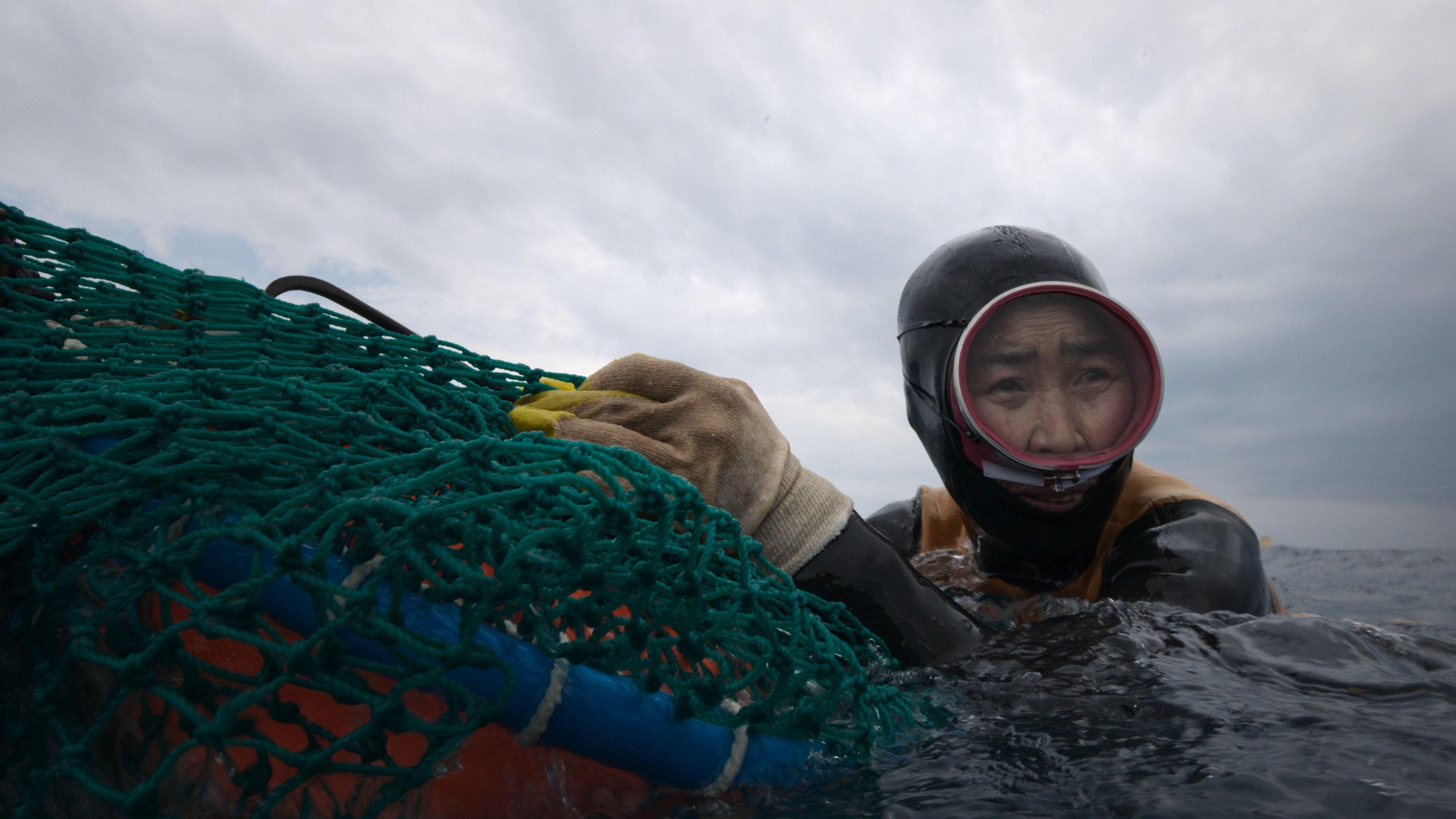 A haenyeo diver from South Korea’s Jeju Island in The Last of the Sea Women, wearing protective yellow gloves and diving gear as she holds on to a green fishing net