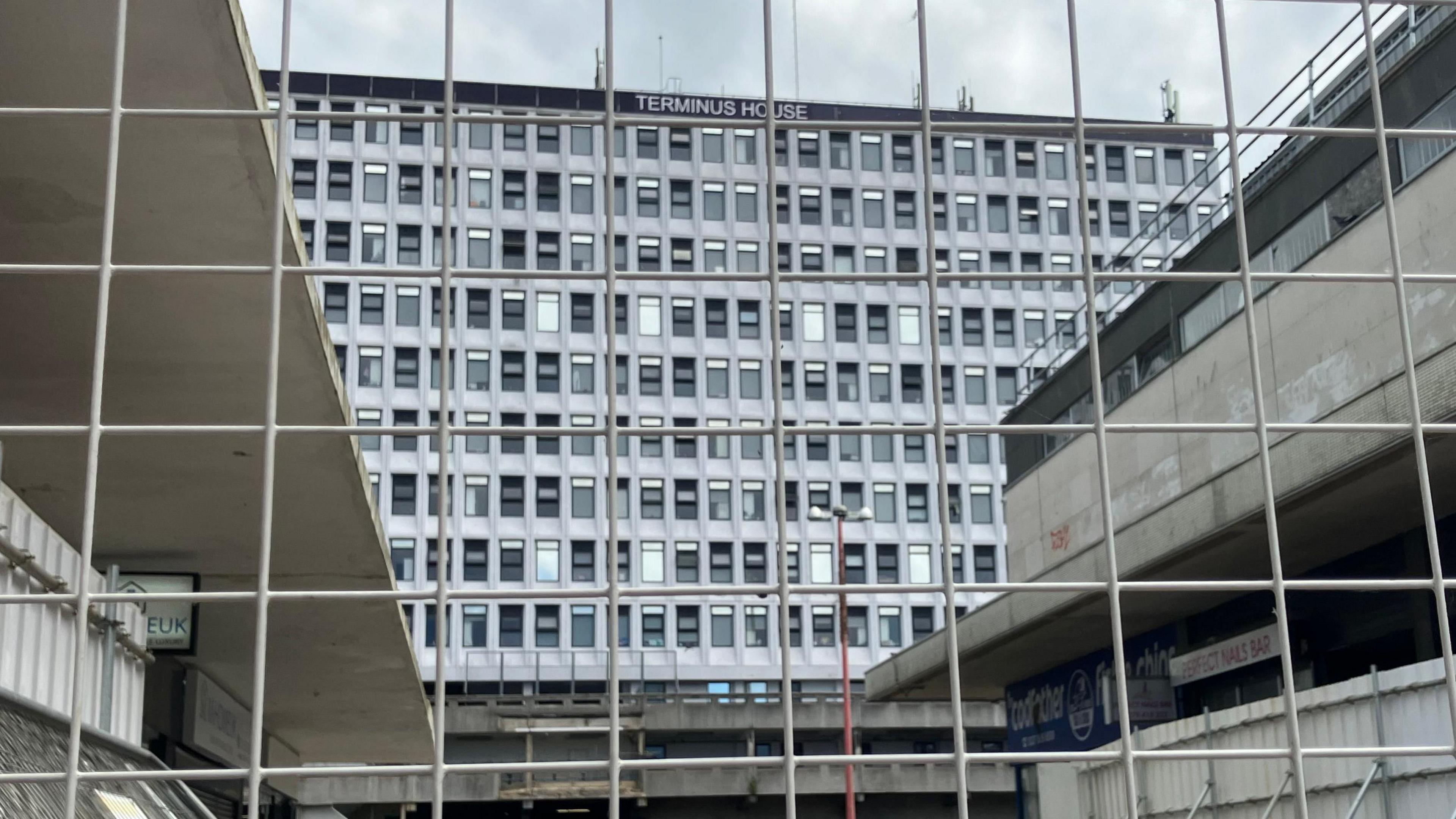 Terminus House in Harlow, seen through a large metal fence. It is an imposing silver building, formerly an office block, with lots of windows spread across it.