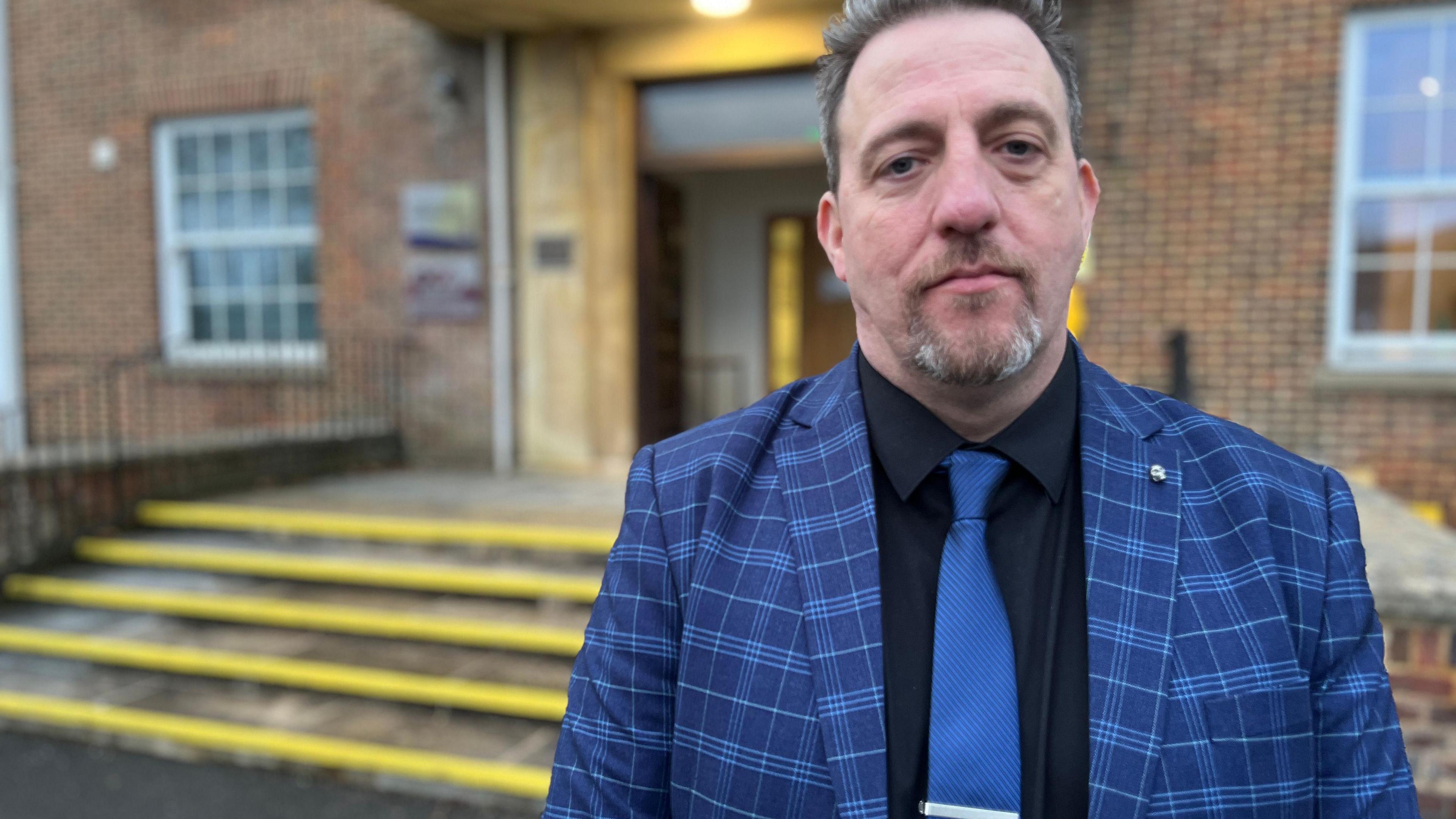 Phil Matthews-Dawson outside Devizes Police Headquarters. He is wearing a checked blue jacket, a dark shirt and a bright blue tie. He has a goatee beard and dark hair